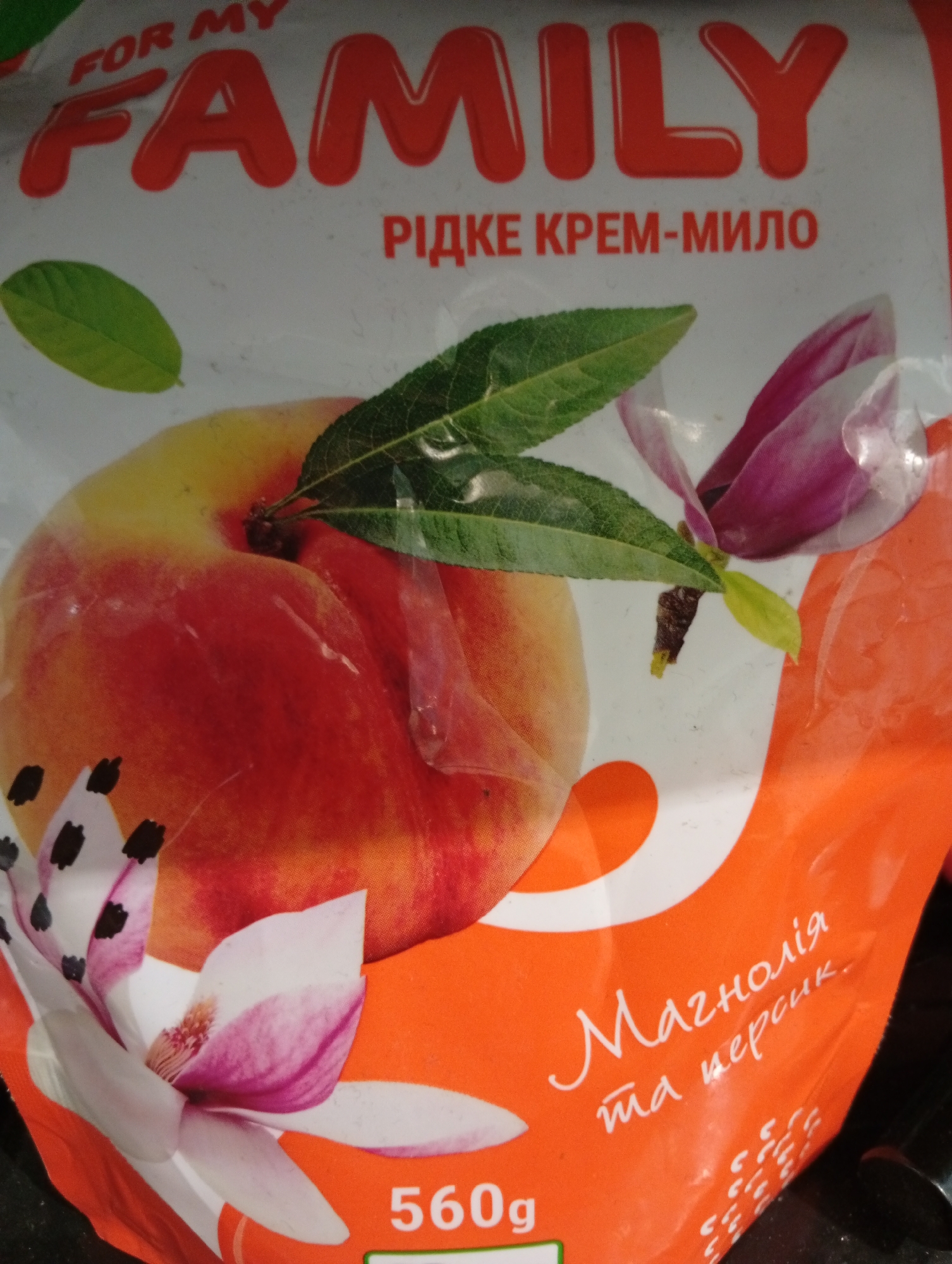 This is a close-up, eye-level, indoor shot of the packaging for a liquid cream soap product. The packaging is predominantly white and orange, featuring graphics of a peach, magnolia flowers, and green leaves. Prominently displayed are the words "FOR MY FAMILY" in large red bubble letters at the top, followed by "РІДКЕ КРЕМ-МИЛО" (which translates to "LIQUID CREAM SOAP") in smaller red text. The bottom right corner of the packaging has white cursive text that reads "Магнолія та персик" (which translates to "Magnolia and peach") and the number "560g" in white text with a green outline. The lighting is artificial and somewhat harsh, casting some shadows and reflections on the plastic packaging.