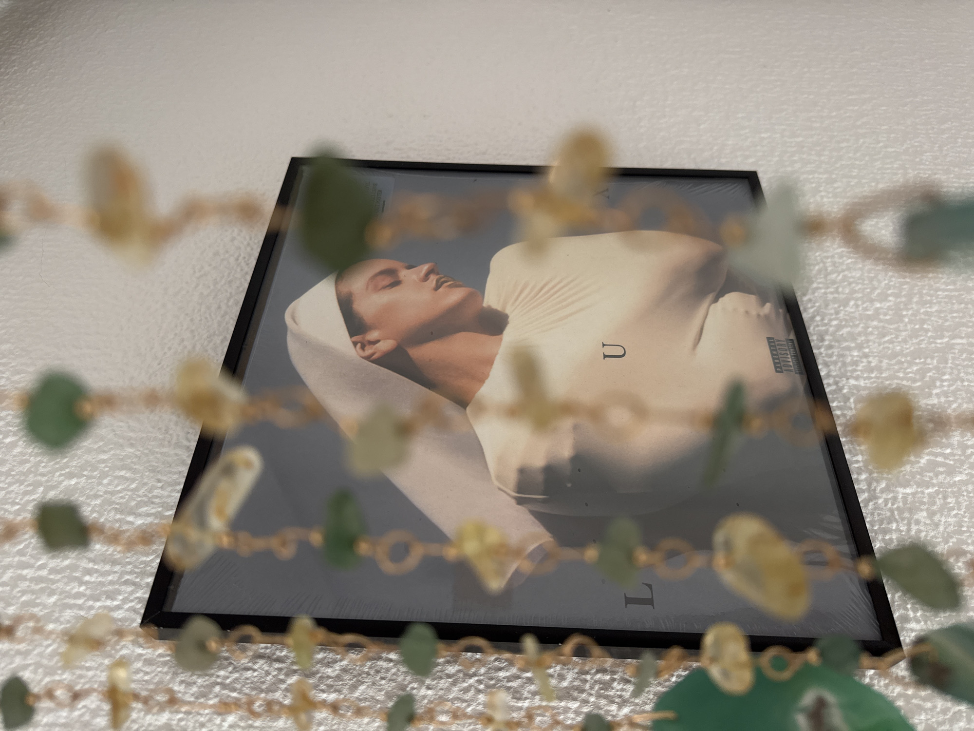 The image shows a framed album cover featuring a close-up of a woman with her eyes closed, resting her head on a cream-colored fabric. She is wearing a white headscarf that drapes over her shoulder. The album cover itself is framed in black and appears to be displayed against a textured white wall. In the foreground, out of focus, is a beaded strand with green and yellow beads, adding a decorative element to the scene. The album cover has text visible, including "PARENTAL ADVISORY EXPLICIT CONTENT" in the bottom right corner. Letters "U" and "L" are also visible on the album cover. The lighting suggests an indoor setting, possibly during the day. The overall mood is serene and artistic. The setting is identified as Las Rozas de Madrid, Spain.