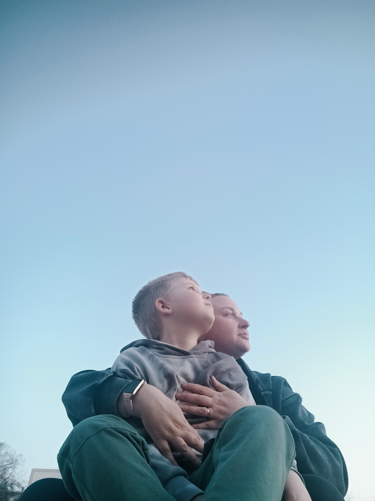 A medium, upward-angled shot depicts a child, assumedly male, cradled in the lap of an adult, assumedly female. Both figures are gazing upward, their faces turned towards the upper right corner of the frame. The child, with light blond hair, is wearing a gray hoodie. The adult, with shorter hair, is wearing a dark jacket. Their hands are intertwined over the child's chest. The background is a pale blue sky. A partial view of a tree with no leaves and a white, nondescript building are visible at the bottom left of the frame. The location, Chaikovsky, Russia, is not discernible from the visual information provided.