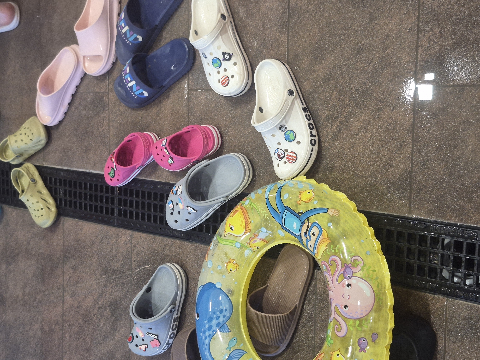 The image shows a collection of footwear and a yellow inflatable swim ring scattered on a brown tiled floor, adjacent to a black metal drainage grate. The footwear consists of several pairs of Crocs-style shoes in various colors (pink, blue, white, gray, and olive green) and sizes, along with a pair of brown slide sandals nestled inside the swim ring. The swim ring features underwater-themed cartoon graphics, including a diver, various fish, and an octopus. The scene suggests a wet environment, possibly a poolside area or a changing room.
There is no information or indication about the location of the city of Votkinsk, Russia.