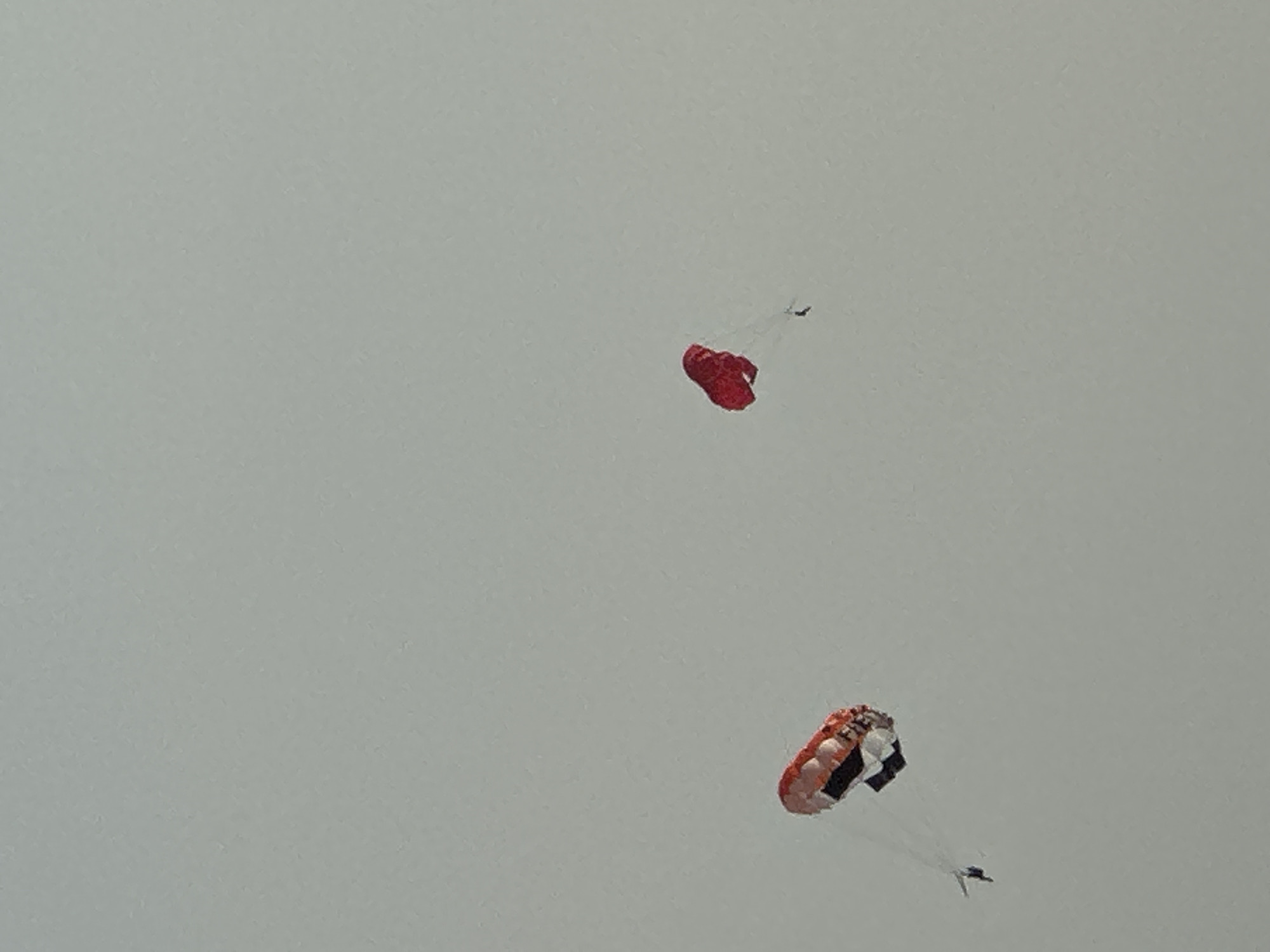In Boreti, Montenegro, two paragliders are captured against a muted, overcast sky. The paraglider on the left features a canopy with red, white, and black sections, with a person suspended below. To the right, a second paraglider displays a solid red canopy, also with a person attached. The figures appear small against the vast expanse of the sky, suggesting they are at a significant altitude. The environment is characterized by an even, diffused light due to the cloud cover.