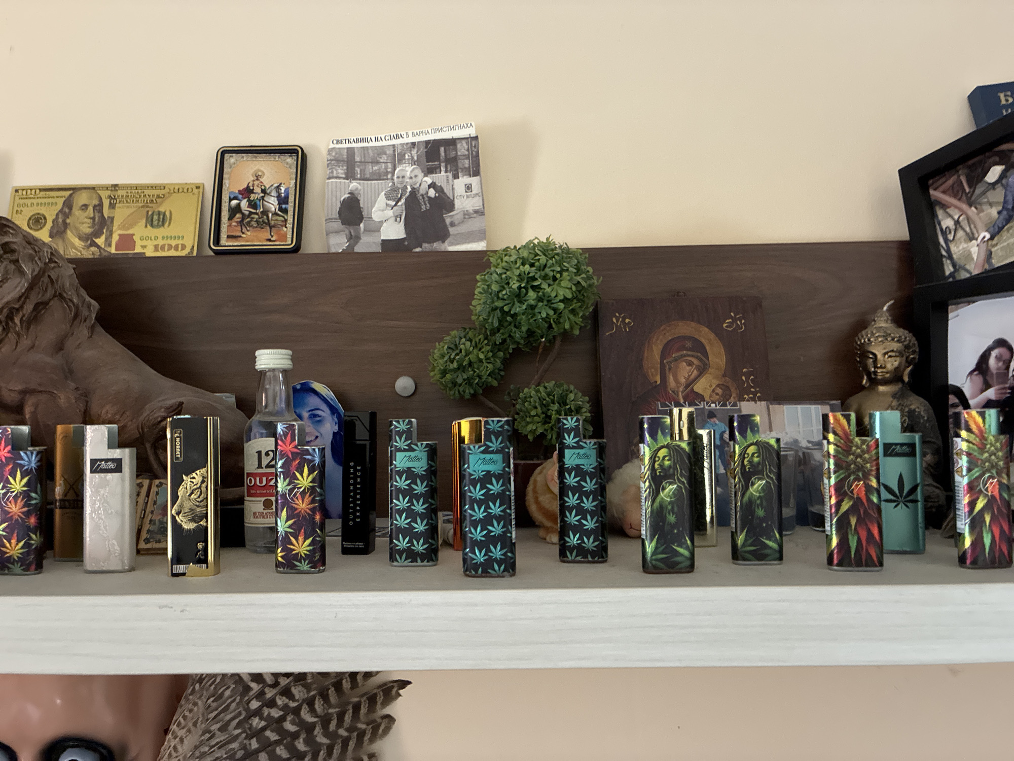 An indoor, eye-level shot captures a diverse collection of personal items displayed on two shelves. The background features a wooden shelf, while the foreground showcases a white shelf below.
On the upper wooden shelf, from left to right, are a gold-colored novelty $100 bill, a small framed religious icon depicting a figure on horseback (possibly St. George), an artificial green topiary plant, a wooden religious icon of the Virgin Mary, and a small golden Buddha statue. To the far left, a large, partially visible carved object, possibly a lion's head, rests. Above these items, on a higher, lighter-colored shelf, are a black and white photograph or book cover titled "СВЕТКАВИЦА НА СЛАВА: В ВАРНА ПРИСТИГНАХА" ("FLASH OF GLORY: ARRIVED IN VARNA"), and various framed photographs.
The prominent feature of the image is the collection of numerous lighters arranged on the lower white shelf. Many lighters feature distinct cannabis leaf designs in various colors (rainbow, teal) or portraits of Bob Marley. Other lighters display a tiger head graphic, or brand names such as "Matteo" and "HORNET". Interspersed among the lighters are a small bottle with text "12 OUZ" (likely Ouzo, possibly Metaxa as suggested by "МЕТАХАСА" on the label), and a black vape device, possibly a Juul. One lighter also features "OUTSTANDING EXPERIENCE" text.
The collection suggests the personal interests of an individual, with themes ranging from spirituality and pop culture to cannabis culture. The visible text on the photo/book, "В ВАРНА ПРИСТИГНАХА", specifically references Varna, Bulgaria.