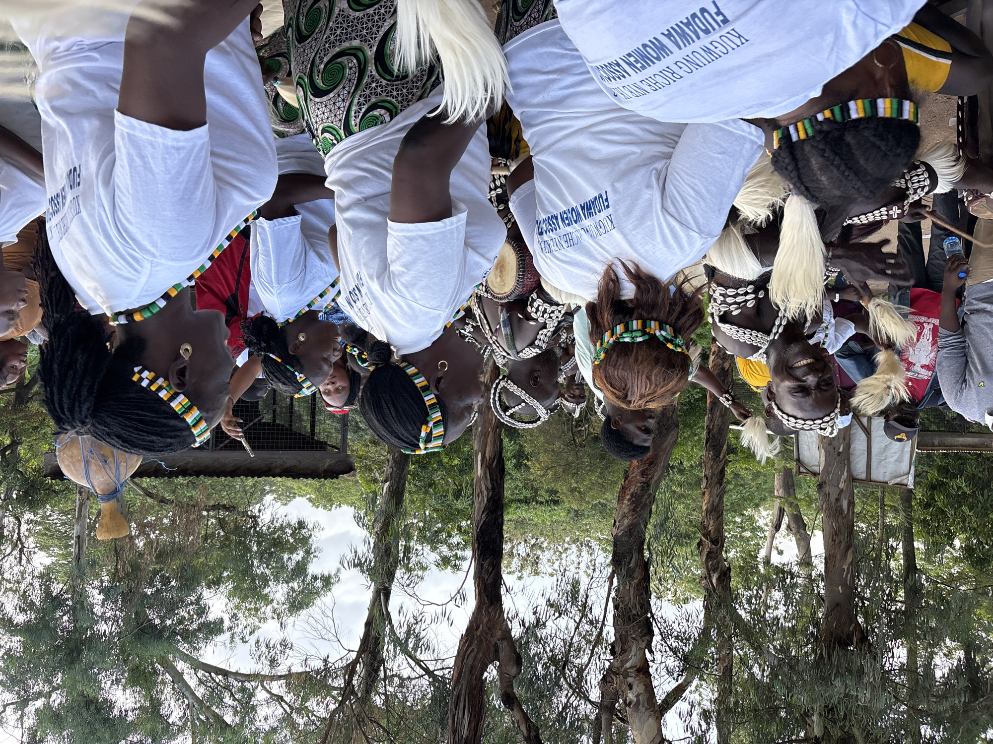 This media file captures a group of people, likely women, gathered outdoors amidst trees. Several individuals are wearing white t-shirts with blue text that reads "FUDAWA WOMEN ASSOC KUGWUNG RICHE NYE". Some are adorned with traditional beaded headbands and necklaces made of shells and beads in green, yellow, and white. One person is wearing a patterned fabric draped over their shoulders.
The scene appears to be part of a cultural event or gathering. There is a drum visible, a water bottle is being held by one person, and another holds a thin stick. The people are looking in various directions, and some appear to be smiling, suggesting a positive and engaging atmosphere. The presence of trees and natural foliage indicates an outdoor setting, possibly a park or a rural area. The lighting suggests it might be daytime, and the sky is cloudy. There is a visible structure in the background, possibly a stall or shelter. The text on the t-shirts suggests the group is affiliated with the "Fudawa Women Association".