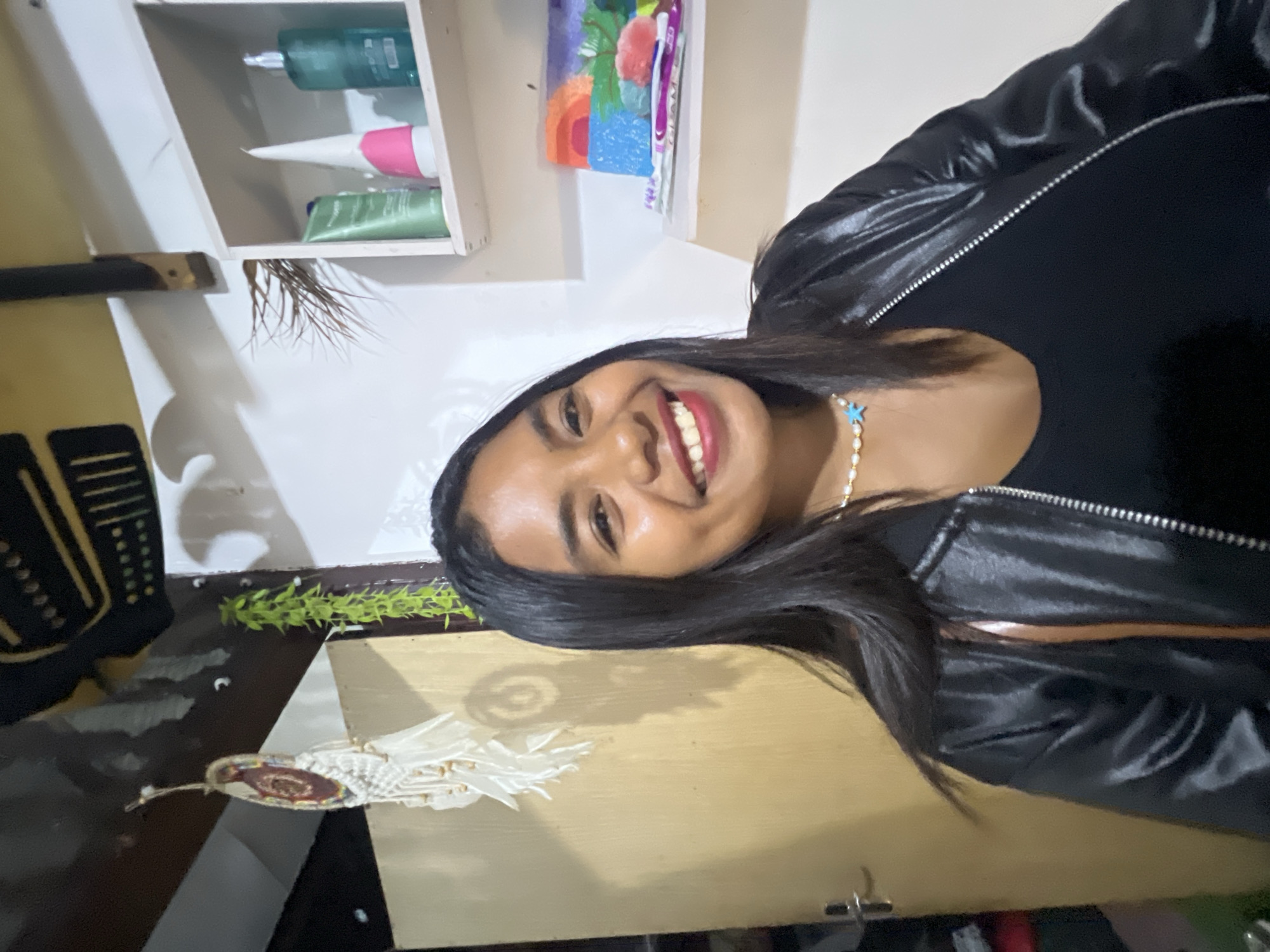 In a domestic interior space, possibly in Denpasar, Indonesia, a young woman with long, dark hair smiles widely at the camera. She is wearing a black top and a shiny black jacket. Around her neck, she has a pearl necklace with a blue starfish pendant. To her left, a tan-colored door with a silver handle is visible, and a dreamcatcher hangs from the ceiling. To her right, a white shelf unit holds several items, including toiletries and a colorful, child-like drawing of a landscape. The background appears to be a wall, with some decorative elements and possibly other rooms visible in the blurred background. The lighting suggests an indoor setting, likely during the day.