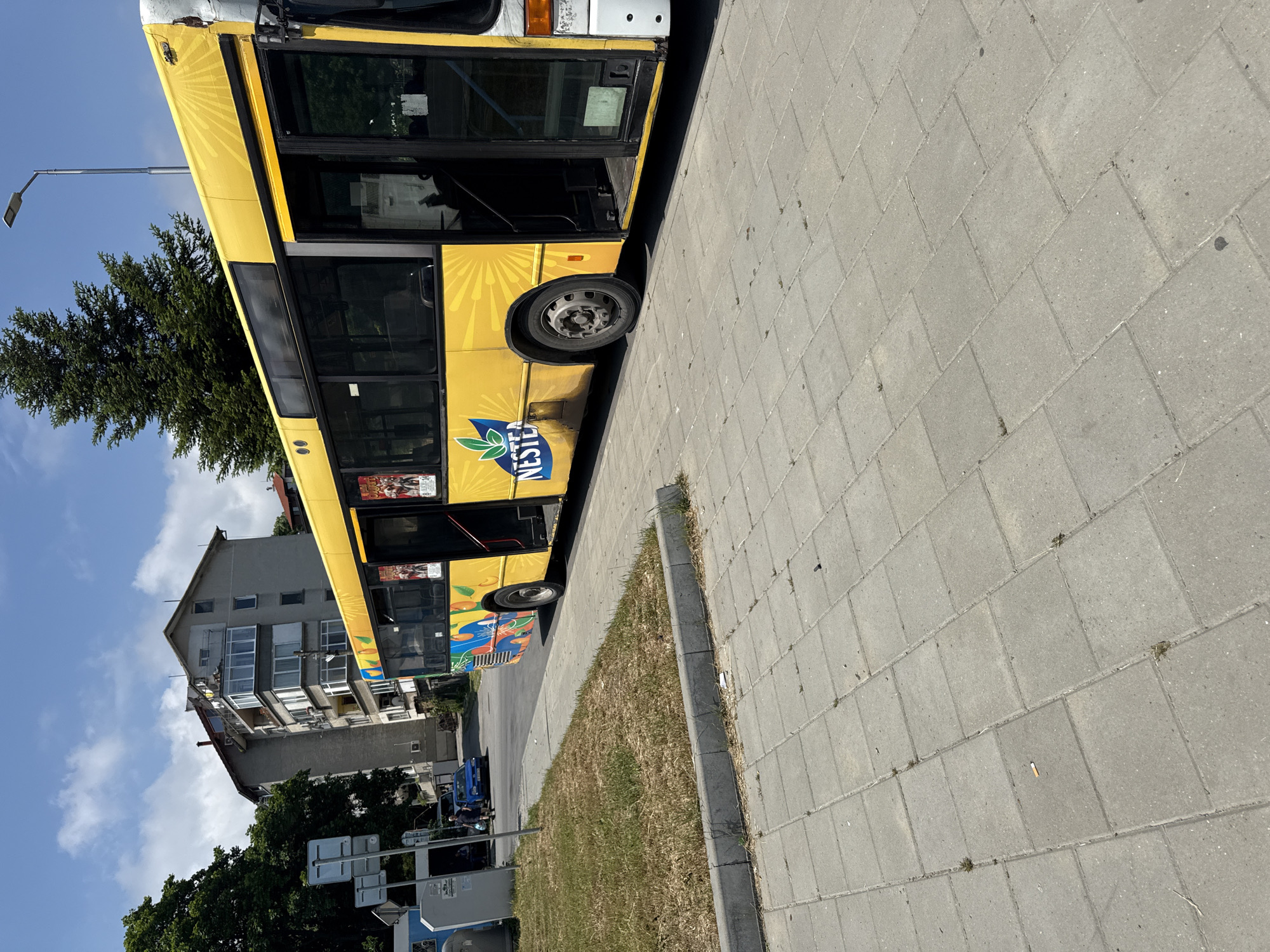 An outdoor, eye-level shot captures a sunny urban street scene featuring a yellow bus parked alongside a paved sidewalk. The image is bright, with a clear blue sky and scattered white clouds overhead, suggesting a daytime setting, likely morning or afternoon.
The main subject is a long, yellow public transport bus, facing the left side of the frame. Its front door is open, revealing a dark interior. The bus features a distinct yellow sunburst graphic along its side and the prominent "NESTLÉ" brand name with its green leaf logo. Below the windows, there are other colorful, abstract designs. The front of the bus shows some signs of wear or minor damage near the roofline.
To the right of the bus, a grey sidewalk made of rectangular pavers stretches into the foreground. Sparse patches of weeds grow in the cracks, and a discarded cigarette butt is visible on the pavement. Between the bus and the sidewalk, a narrow strip of grass, mostly dry and brown, runs parallel to the street.
In the background, behind the bus, a multi-story residential building with multiple windows and balconies is visible under the blue sky. Green trees are interspersed throughout the scene, providing natural elements. A tall, grey street lamp post extends upwards from the left, against the sky. Further down the street, beyond the bus, a smaller blue vehicle is partially visible, along with some road signs.
No people are clearly visible, and there is no overt activity taking place, suggesting a moment of pause, perhaps at a bus stop. The overall impression is one of a typical, active urban environment on a clear day.