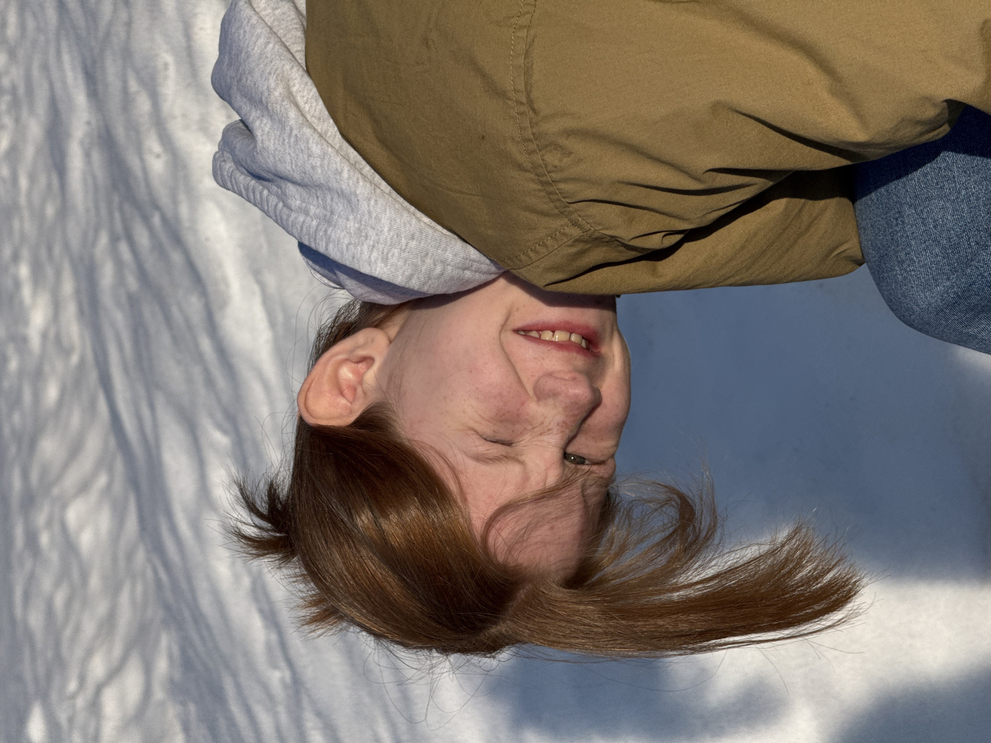 The image depicts a close-up, head-and-shoulders view of a young individual positioned outdoors in a snowy environment in Chaikovsky, Russia. The subject, with reddish-brown hair and fair, freckled skin, has their head tilted approximately 45 degrees to the left (from the viewer's perspective). The left eye is partially closed or squinted, and the mouth is open, revealing upper and lower teeth, with the corners pulled back in a pronounced expression. The lips exhibit a reddish tint. The individual is wearing a light grey hooded sweatshirt under an olive-drab or khaki-colored jacket, which features visible stitching details on the right shoulder and upper back. A section of blue denim fabric is visible at the lower right of the frame. The background consists entirely of snow, showing undulating textures and distinct, elongated bluish shadows, indicative of strong, direct sunlight. No other individuals or objects are discernible within the frame.