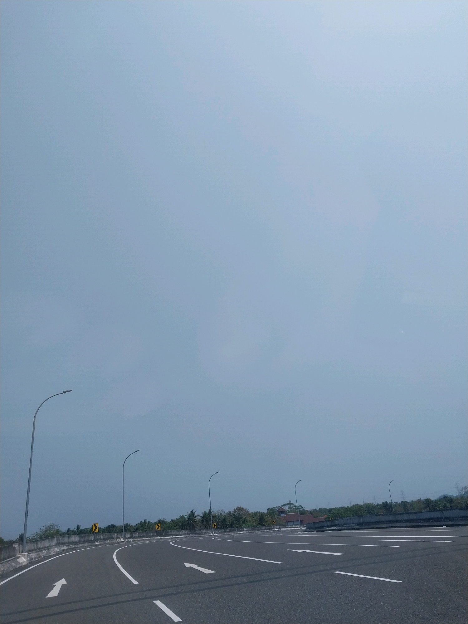The image captures a view of a highway in Palihan, Indonesia, under an overcast sky. The road is paved and marked with white lane lines, including directional arrows. Several streetlights are visible along the side of the road, and yellow warning signs indicating curves are posted. A concrete barrier separates the highway from the surrounding landscape, which includes trees and some buildings in the distance. The overall scene suggests a typical day on a well-maintained road, with no people or vehicles immediately visible.