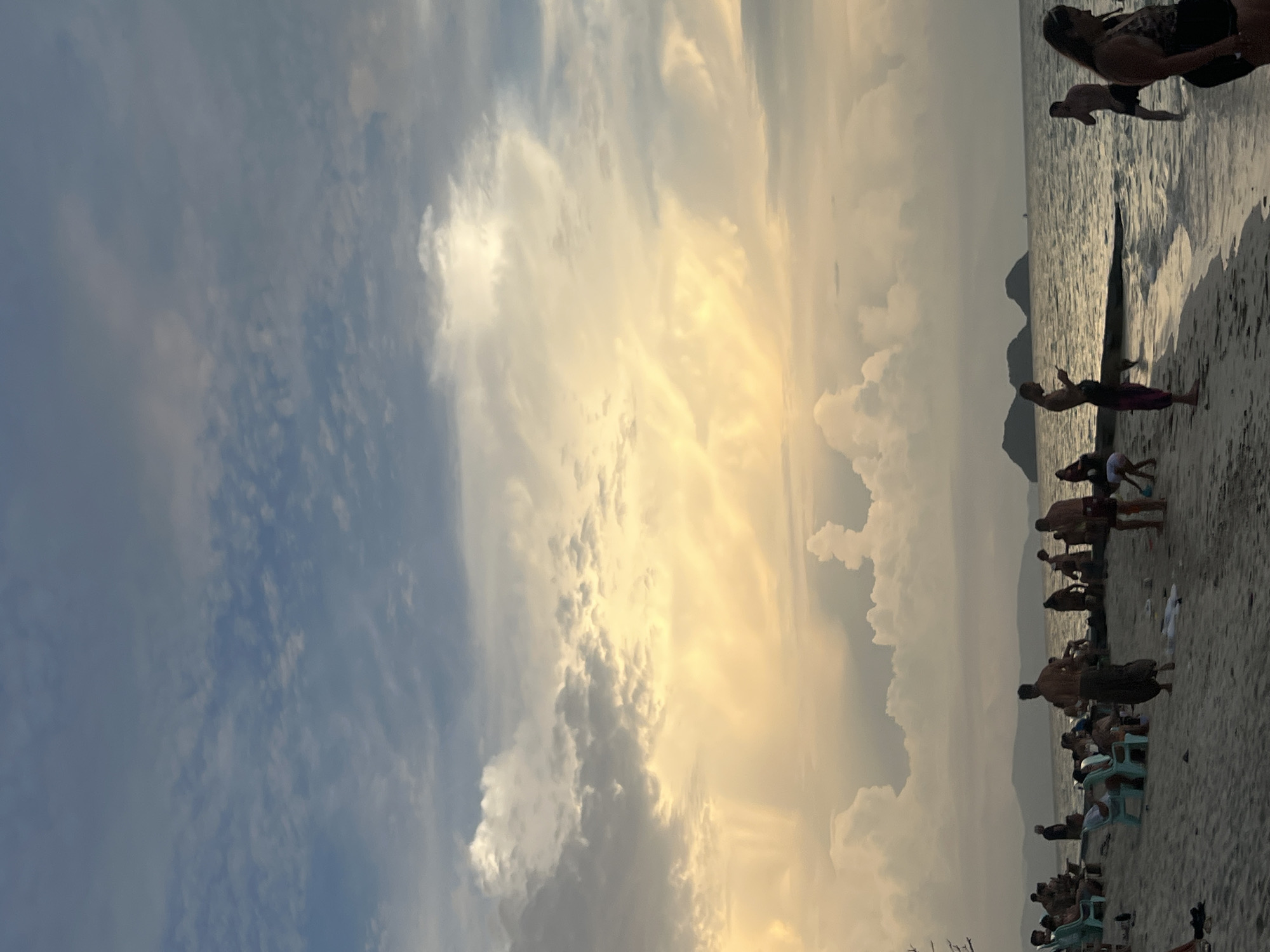 This image depicts a beach scene on the island of Ko Pha-ngan, Thailand, during what appears to be sunset or dusk. The sky is a prominent feature, filled with dramatic clouds illuminated by warm, golden light, contrasting with areas of blue and gray. The ocean is calm, with gentle waves lapping at the shore.
On the sandy beach, several people are visible. Some are seated on chairs, while others are standing or walking along the shoreline. A few individuals are in the shallow water. The overall atmosphere suggests a relaxed, communal gathering or a leisurely evening on the beach. In the background, distant islands or hills are silhouetted against the horizon.
