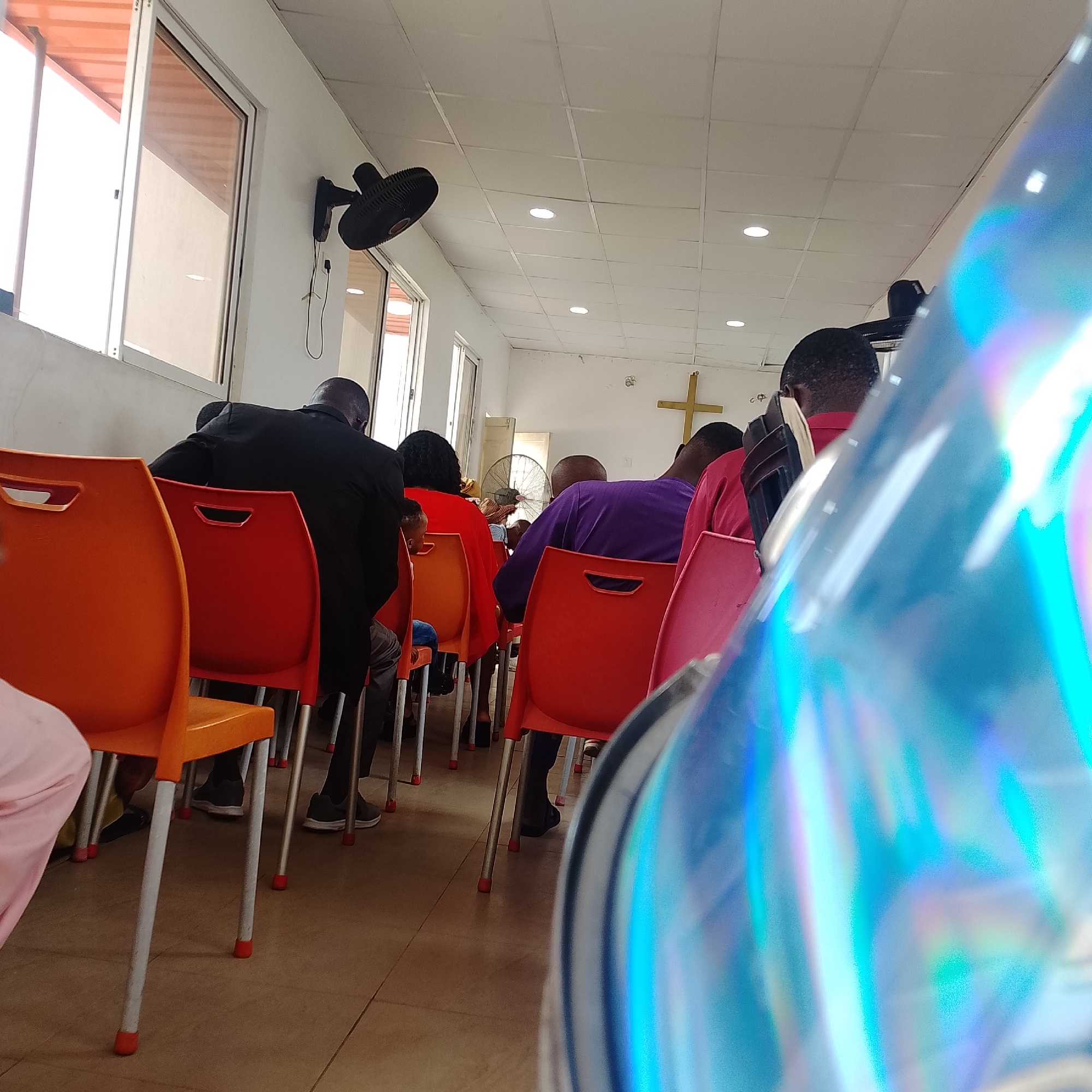 This image depicts the interior of a church in Iranla, Nigeria, with a focus on the congregation seated on colorful plastic chairs. In the foreground, a blurry, iridescent surface, likely a camera lens or a reflective object, dominates the lower right portion of the frame. The background reveals a white-walled room with a white tiled ceiling, illuminated by recessed lighting. A black oscillating fan is mounted on the left wall, and a large, golden cross is visible on the far wall. The congregation consists of men and women of various ages, dressed in church attire. Some are facing forward, while others are looking away from the camera. The chairs are arranged in rows, with some occupied and others empty. The overall atmosphere suggests a religious service is in progress or about to begin.
