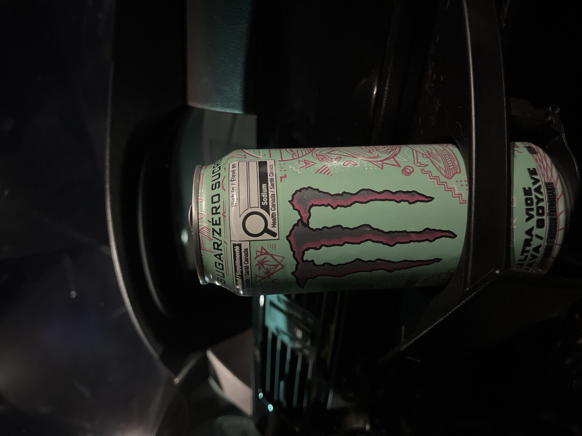 A mint-green can of Monster Ultra Vice energy drink is wedged inside a black plastic car cupholder. The can's label indicates "SUGAR/ZÉRO SUCR" and "ULTRA VICE AVA / GOYAVE." Part of a car dashboard is visible around the cupholder. The image was not taken in Shumen, Bulgaria, and there is no evidence to suggest it was.