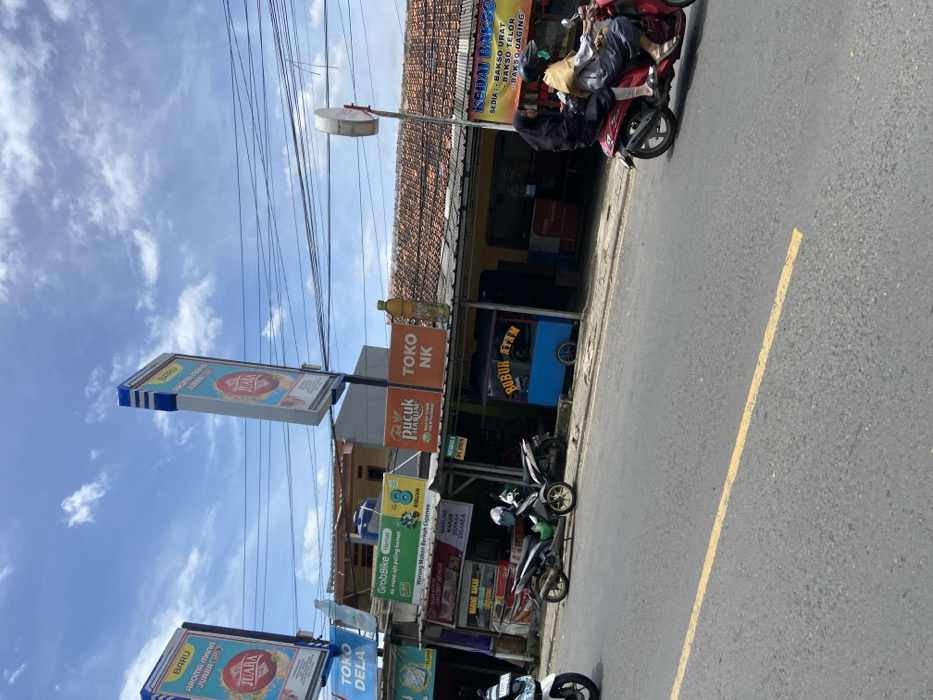 The image depicts a street scene in Jatiluhur, Indonesia (latitude: -6.565, longitude: 107.434). Two women are riding a red motorbike down the paved street, a yellow line down the center. In the background, there are several shops and stalls, including a "Bubur Ayam" (chicken porridge) stall, a "Toko NK" shop, and a "Kedai Bakso" (meatball stall). The street is lined with power lines. There is a sign for "Aroma Teh Minuman Juara" (Champion Tea Drink). The sky is blue with white clouds. The image captures a typical street scene in Jatiluhur, Indonesia, featuring everyday life, local businesses, and the ubiquitous motorbike transportation.