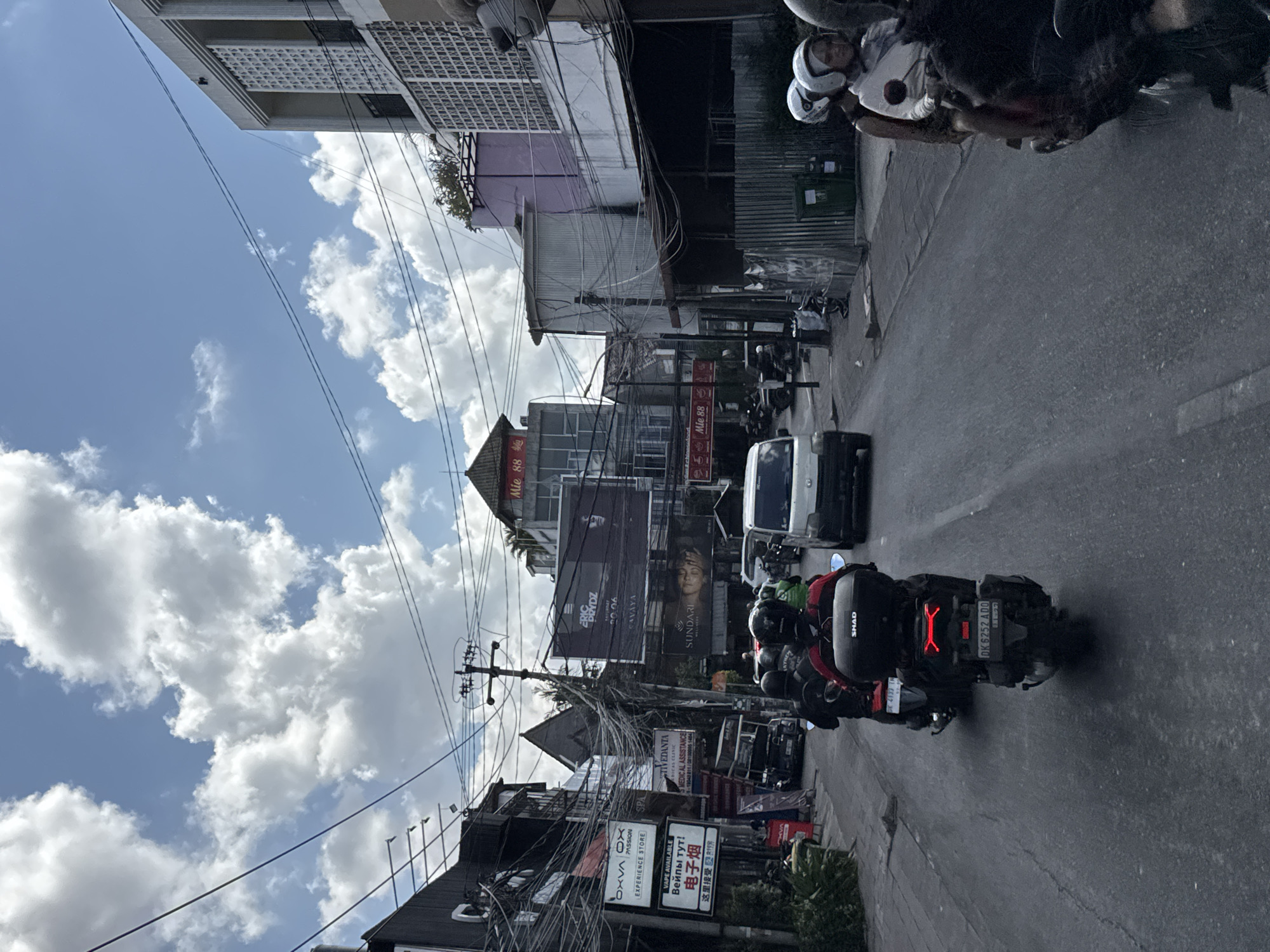 This upright image depicts a bustling street scene in North Kuta, Indonesia, under a bright, cloudy sky. The perspective is slightly elevated, looking down an asphalt road flanked by various buildings. A dense, intricate network of overhead power lines and utility cables dominates the view above the street.
In the foreground, a dark motorcycle with a prominent "SHAD" brand top case and a distinctive red X-shaped taillight is a central subject. Its rider, wearing a red jacket and helmet, is visible. The Indonesian license plate "DK 6252 ADO" is clearly legible on the motorcycle, with "DK" indicating Bali. Further down the road, other motorcycles and a white van suggest active traffic.
The buildings lining the street are a mix of commercial establishments and multi-story structures, some with balconies featuring decorative grilles. Numerous commercial signs are affixed to these buildings and utility poles. Visible text includes "Mie 88 麵" (a noodle shop, appearing multiple times), "OXVA OX PASSION EXPERIENCE STORE," and multilingual signage for vaping products: "VAPE AVAILABLE," "Вейпы тут!" (Russian), and "电子烟," "这里接受" (Chinese). Additional signs advertise "SPA," "STIVEDANTA MEDICAL CLINIC," "MEDICAL ASSISTANCE," "CAVAYA," and "SUNDARI."
The weather is sunny with scattered white cumulus clouds against a blue sky, indicating midday or early afternoon. The scene captures the lively, cluttered character typical of urban streets in Southeast Asia, with a constant flow of activity.