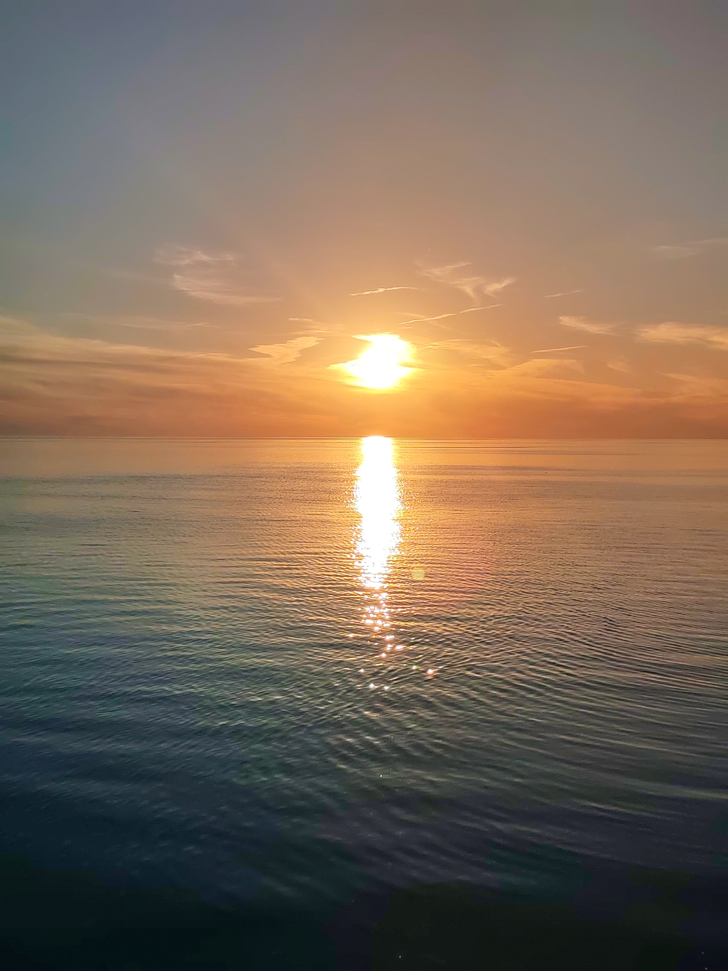 The image depicts a tranquil sunset over a vast body of water, with no land visible on the horizon. The sky transitions from a pale blue-grey at the top to vibrant shades of orange, peach, and gold near the horizon line. Sparse, elongated cirrus clouds and what appear to be aircraft contrails are scattered across the upper and middle sky. The sun, a brilliant white-yellow orb, is positioned slightly above the horizon, off-center to the right, casting an intense, shimmering vertical reflection across the water's surface that extends towards the foreground. The water itself displays a gradient of colors, mirroring the sky with warm golden and orange tones near the horizon, gradually shifting to deeper blues and greens in the foreground. The surface of the water is characterized by numerous small, undulating ripples that catch and scatter the sunlight, creating sparkling highlights, particularly within the sun's reflection path. No individuals, vessels, or man-made structures are present within the frame. The scene conveys an open, natural marine or lacustrine environment.