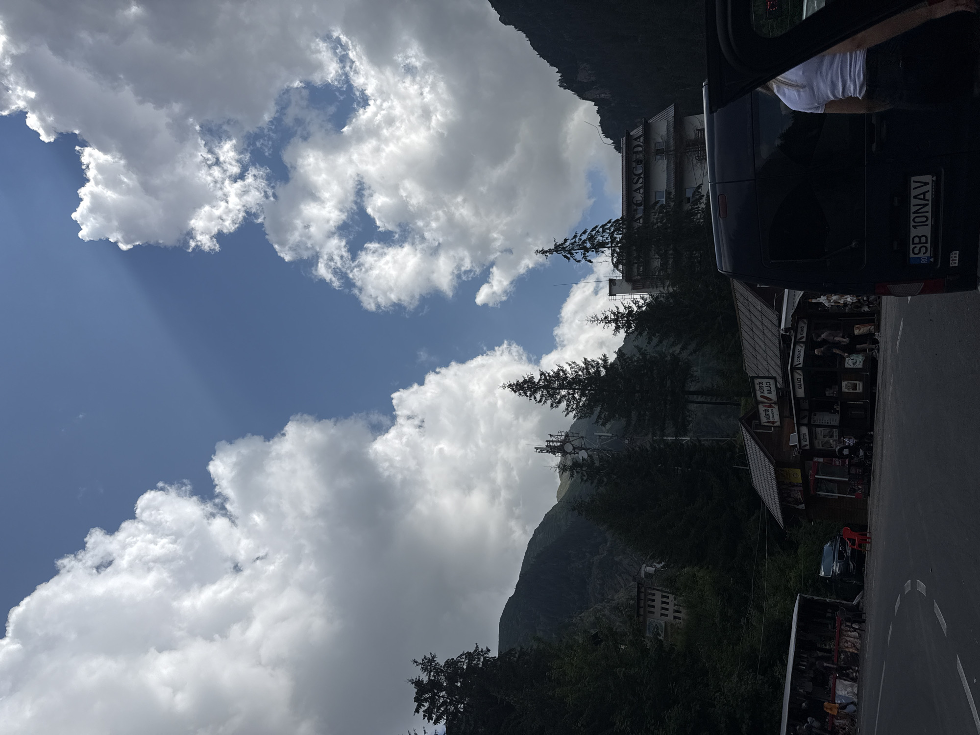 The image depicts a scenic view of a mountainous area with a building labeled "A CASCADA" in Cârțișoara, Romania. The sky is partly cloudy with dramatic light rays piercing through the clouds. In the foreground, there are several small structures that appear to be shops or stalls, one of which has signage for "KÜRTŐS KALACS." A black van is parked on the right side of the road, with its side door open. A person wearing a white t-shirt and dark shorts is visible standing near the open door. A red plastic chair is placed on the asphalt in front of one of the stalls. The road curves to the left, and a black car is parked further down. Tall evergreen trees flank the scene, and in the background, a large, multi-story building, potentially a hotel or a significant structure, is partially visible amongst the trees on the mountainside. There are also telecommunication towers visible on the mountain. The overall impression is a roadside stop in a mountainous, possibly touristy, region.