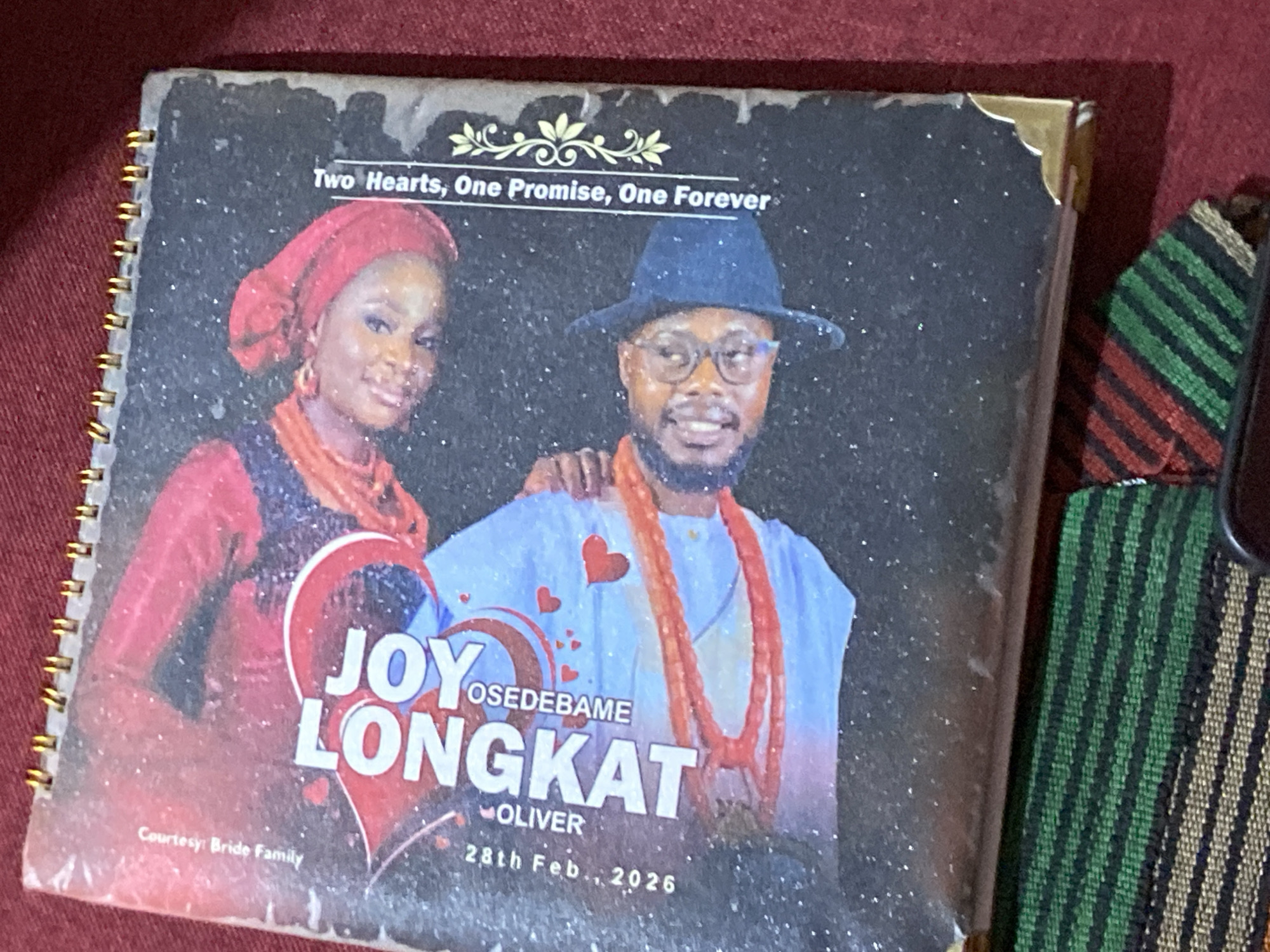 The image is a close-up overhead shot of a spiral-bound notebook lying on a dark red fabric surface. The notebook cover features a glittery black background with an image of a smiling couple, presumably for a wedding or engagement. The couple is dressed in festive attire, with the man wearing a blue hat and the woman wearing a red headscarf. Text on the cover reads "JOY LONGBAT" and "OSEDEBAME OLIVER," along with a date "28th Feb., 2026." The bottom of the cover has the text "Two Hearts, One Promise, One Forever" and a decorative floral motif. A snippet of a colorful striped fabric, possibly part of a garment or decoration, is visible on the left side of the notebook. The notebook has gold spiral binding on the right edge and gold corner protectors on the bottom left. The overall scene suggests a celebratory event. The text "Courtesy Bride Family" is visible in the upper right corner, indicating the origin of the notebook or the event it commemorates. The location is identified as Rukuba Road, Nigeria.