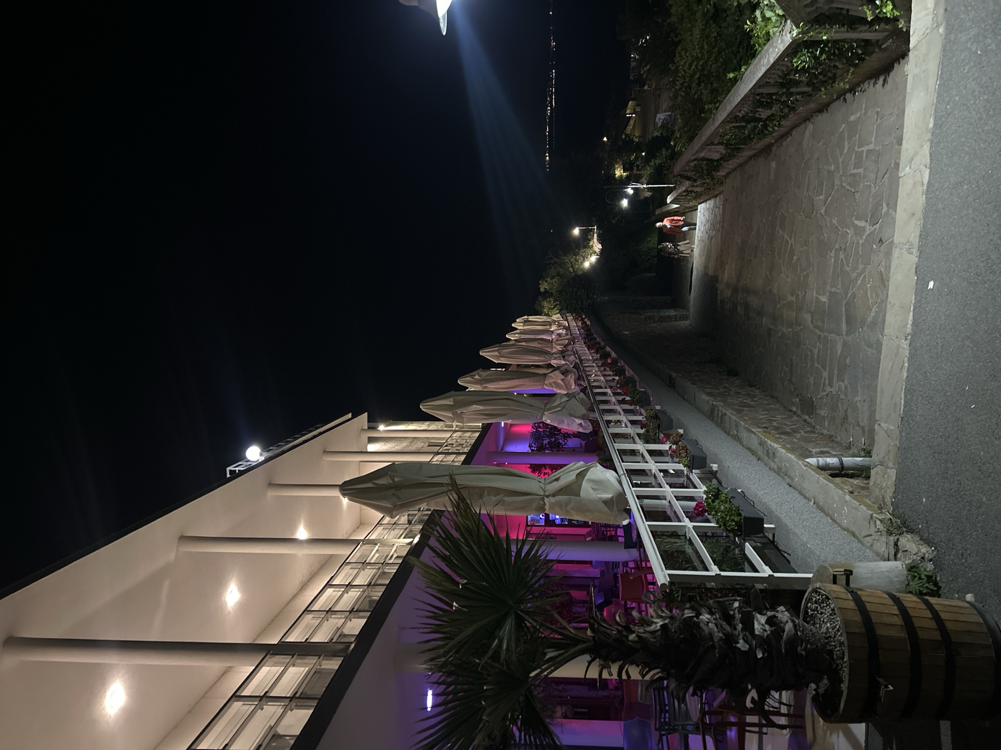 At night, a row of closed, white umbrellas lines the exterior of a white building with large windows. The building has a flat roof with support columns. The interior is lit with vibrant purple and red lights. A palm tree and a wooden barrel planter sit at the front of the building. Adjacent to the building, there's a stone walkway and road. In the distance, city lights are visible. Based on similar images, this may be a restaurant in Burgas, Bulgaria. There are no people visible.