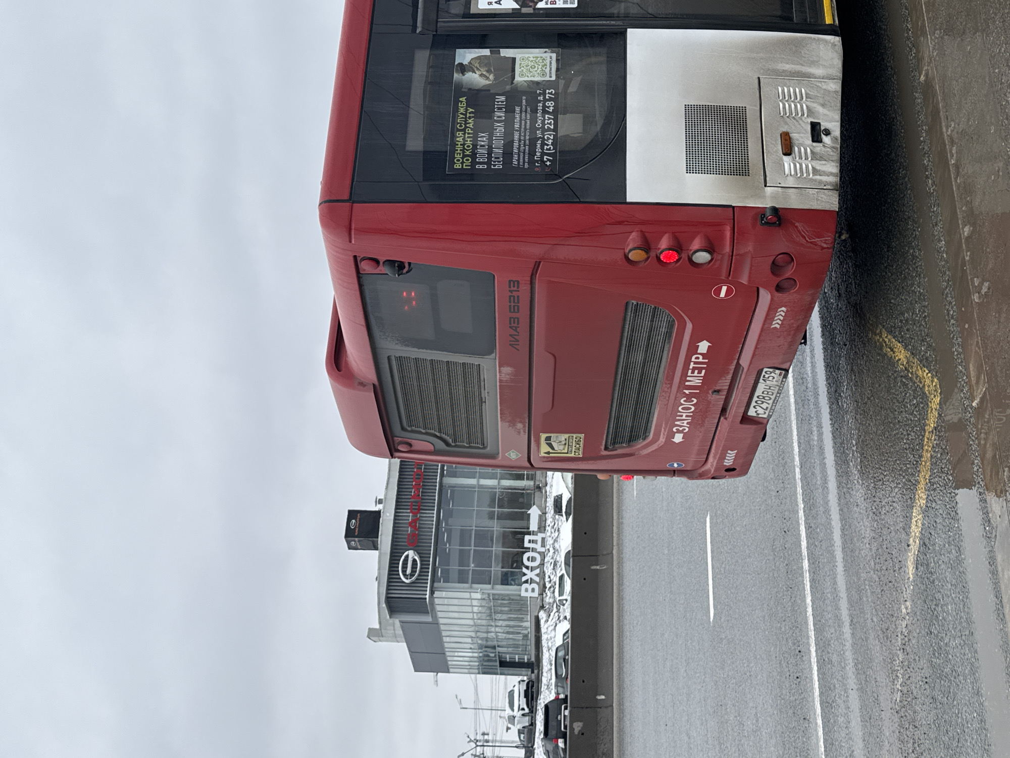 A red and white LiAZ 6213 city bus is depicted from its rear-right side, parked or moving slowly on a wet asphalt road. The road features white lane markings and a yellow line. The sky is overcast and grey, suggesting a cloudy day, and the wet road indicates recent rain or slushy conditions.
The bus displays several notable details. On its side, a prominent advertisement for military service reads: "ВОЕННАЯ СЛУЖБА ПО КОНТРАКТУ В ВОЙСКАХ БЕСПИЛОТНЫХ СИСТЕМ" (Military service by contract in unmanned systems troops). Below this, it states "ГАРАНТИРОВАННОЕ УВОЛЬНЕНИЕ с военной службы по истечении срока контракта при нежелании заключать новый контракт" (Guaranteed discharge from military service upon expiration of the contract if unwilling to sign a new one), along with a Perm address ("г. Пермь, ул. Окулова, д. 7.") and a phone number (+7 (342) 237 48 73). A "ЗАНОС 1 МЕТР" (Swing out 1 meter) warning sign is visible on the rear of the bus, alongside its Russian license plate, "C298BH 159," which includes the Perm Krai region code "159."
To the left of the bus, a modern commercial building with a glass facade is visible, featuring a "BYD" logo and a large sign in front that reads "ВХОД" (Entrance). Several cars are parked in front of this building. No people are distinctly visible in the frame. The overall scene captures an urban environment in Perm, Russia, under gloomy weather conditions.