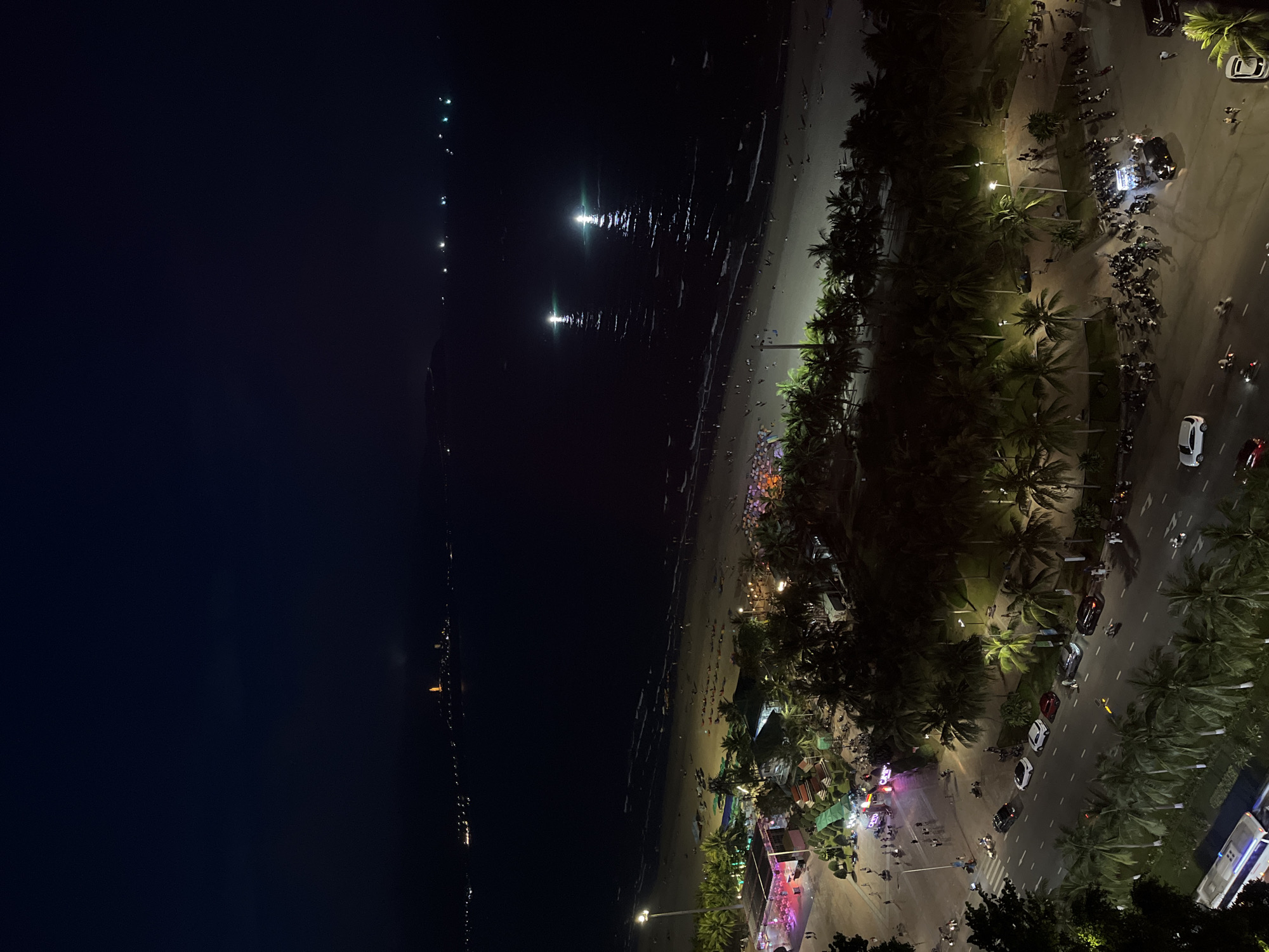 This is a nighttime aerial view of the beach and city life in Đà Nẵng, Vietnam. A dark ocean stretches to the left, illuminated by scattered lights in the distance and two strong spotlights shining onto the water. A sandy beach runs along the shoreline, showing faint signs of people gathered along the lit areas. The beach is bordered by a line of palm trees, beyond which runs a road bustling with motorbikes, cars, and pedestrians. Streetlights illuminate this area. The scene seems lively with activity along the coast and streets.