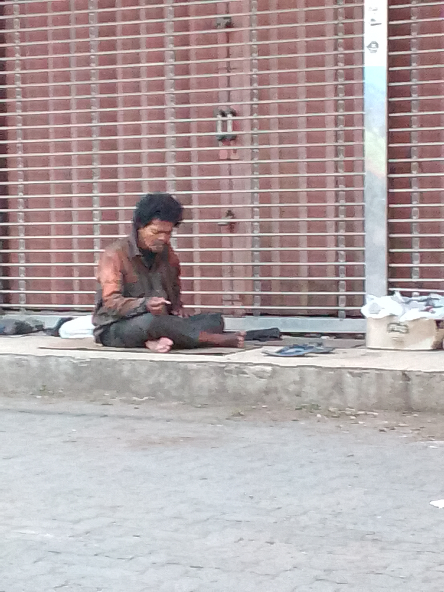 A person sits on a sidewalk in front of a metal corrugated gate. The person is wearing a dark long-sleeved shirt and dark pants. They are sitting cross-legged with their right foot visible. A white pillow is behind them, and there is a piece of cardboard on the ground in front of them. To the right of the person, there is a white bucket filled with what appears to be trash. A pair of blue flip-flops lies on the ground in front of the bucket. The background gate is a reddish-brown color with horizontal white bars. A silver pole is visible on the right side of the frame, with some writing on it. The ground in front of the sidewalk is a paved road.