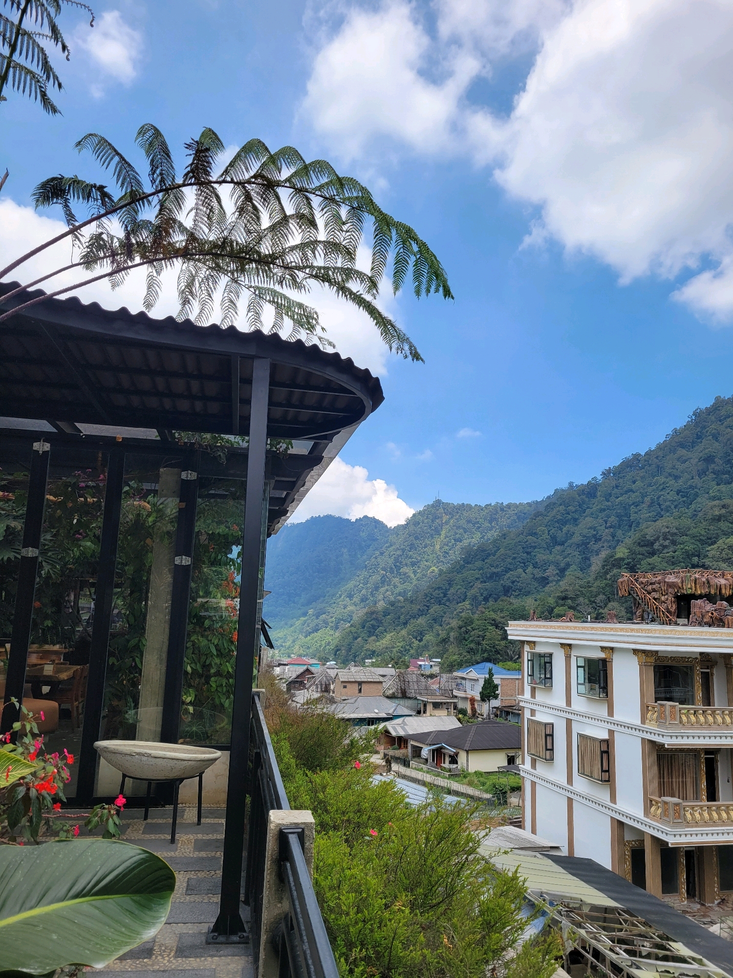 The image captures a scenic view from a balcony overlooking a lush, mountainous landscape in Semangat Gunung, Indonesia. In the foreground, a portion of a modern building with white walls and ornate gold trim is visible on the right. A black metal railing runs along the edge of the balcony, with a large green leaf and red flowers in the lower left corner. Behind the railing, a covered patio area with a black roof and glass walls is present. A stone basin on a stand sits on the patio.
The midground reveals a cluster of houses and buildings with varying roof styles nestled at the base of a steep, densely forested mountain that dominates the background. The sky above is a clear blue with scattered white clouds. The overall impression is of a tranquil, natural setting enhanced by the presence of man-made structures.