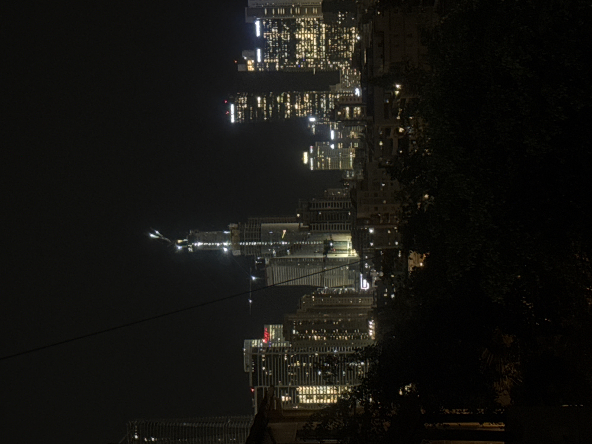Here's a description of the image:
The image is a nighttime view of a city skyline. The location is Givatayim, Israel. The skyscrapers are lit up with many small lights, creating a bright, urban scene that contrasts with the surrounding darkness. Trees and foliage appear in the foreground and right side of the image. A power line crosses in front of the skyline.