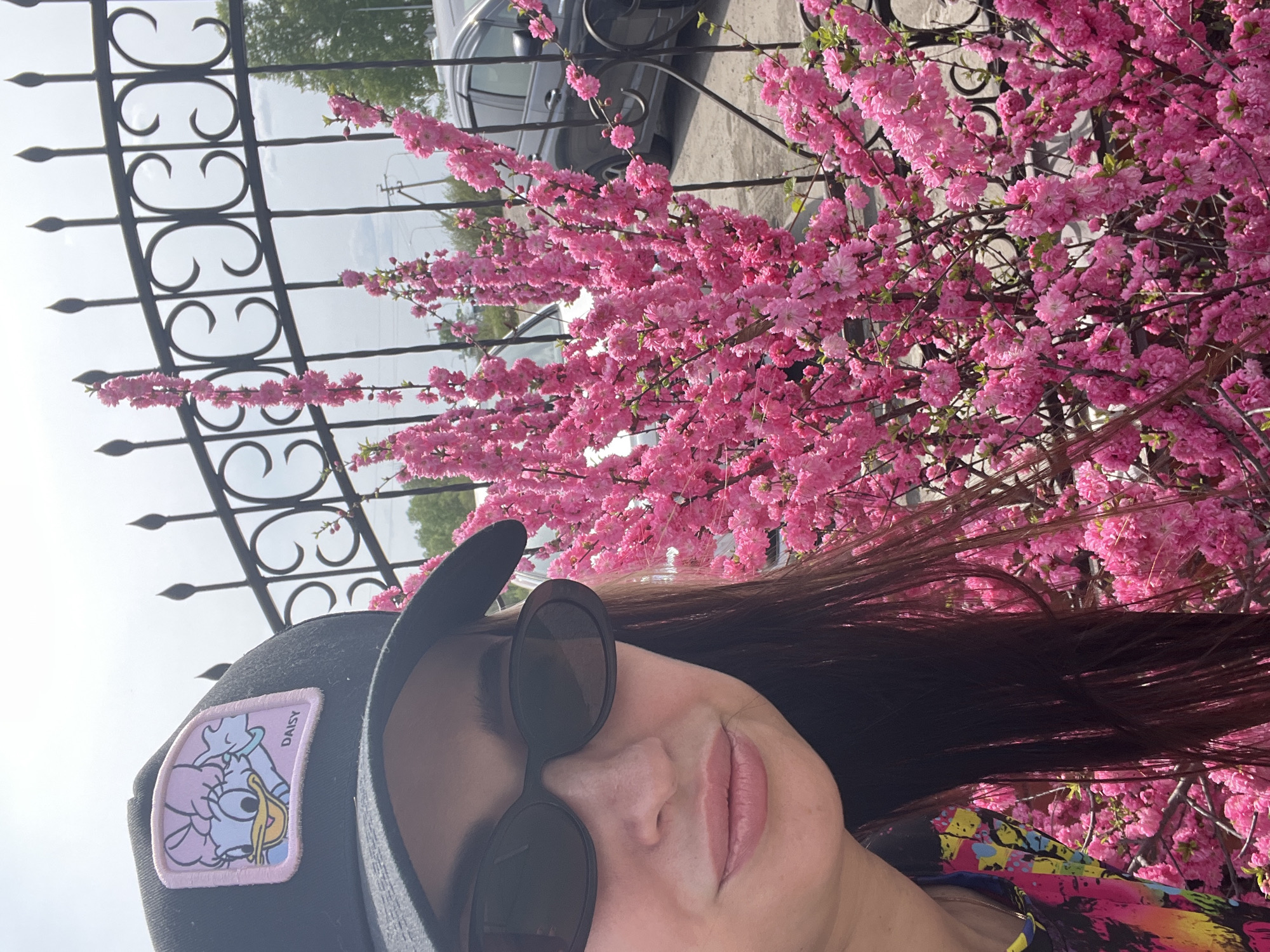 A woman wearing a black baseball cap with a Daisy Duck patch and round sunglasses is photographed outdoors in an urban setting. She is positioned next to a wrought-iron fence and a flowering bush with dense pink blossoms. A dark-colored car is visible in the background, behind the fence and flowers. The location is in the city of Unknown, Unknown.