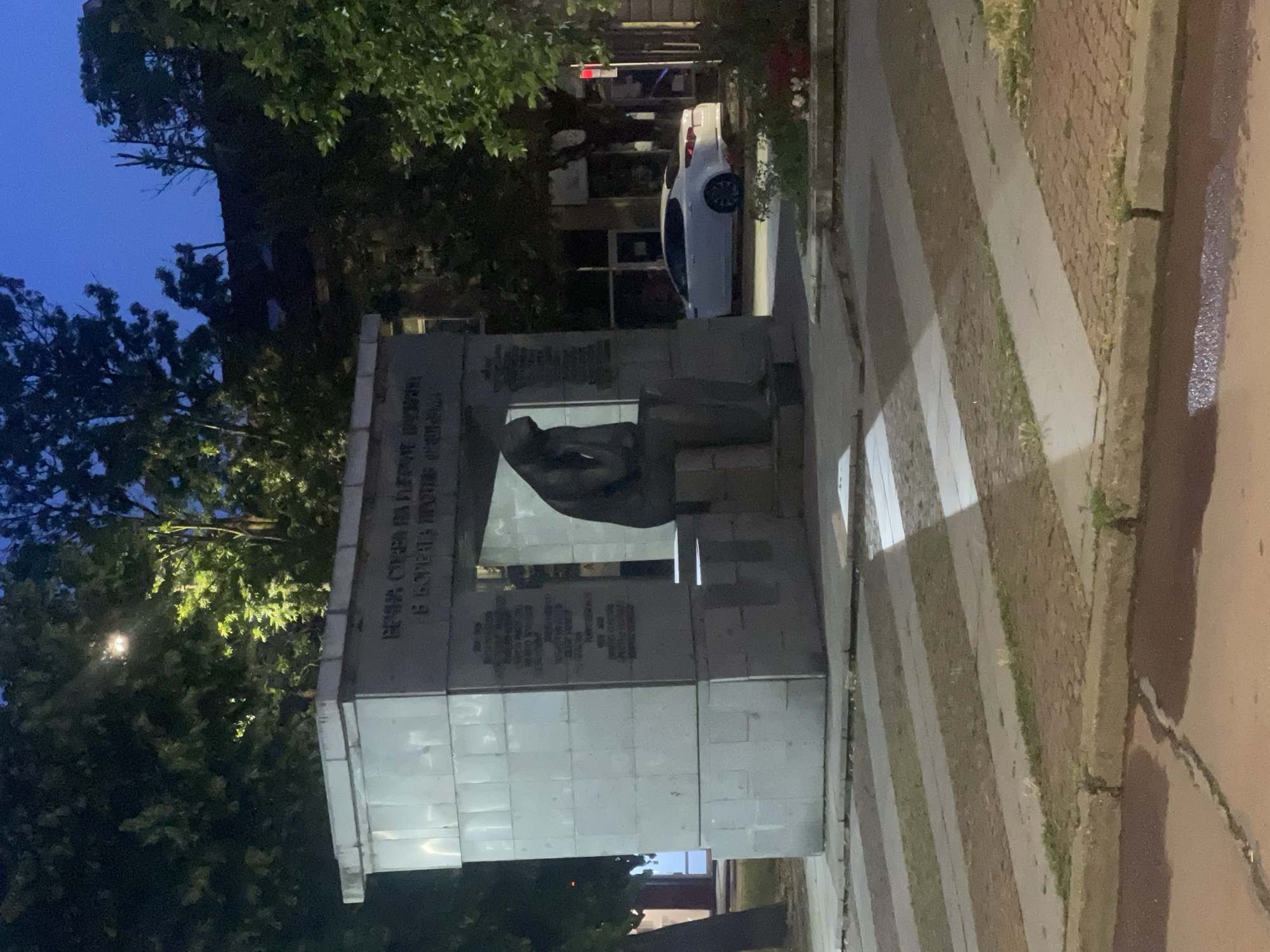 The image depicts a monument in the city of Ruse, Bulgaria, situated in a public square. The monument is a large, rectangular stone structure with an open, recessed area in the center. Within this recess, a seated, cloaked sculpture of a figure is positioned, appearing to be in a contemplative pose. The facade of the monument is adorned with inscribed text. Surrounding the monument is a paved area with a distinct pattern of diagonal stripes of different materials. Trees with dense foliage frame the scene, and a white sedan is parked in the background to the right of the monument. The lighting suggests it is either dusk or early evening, with artificial lights illuminating parts of the monument and the surroundings. The sky is a deep blue, indicating twilight.