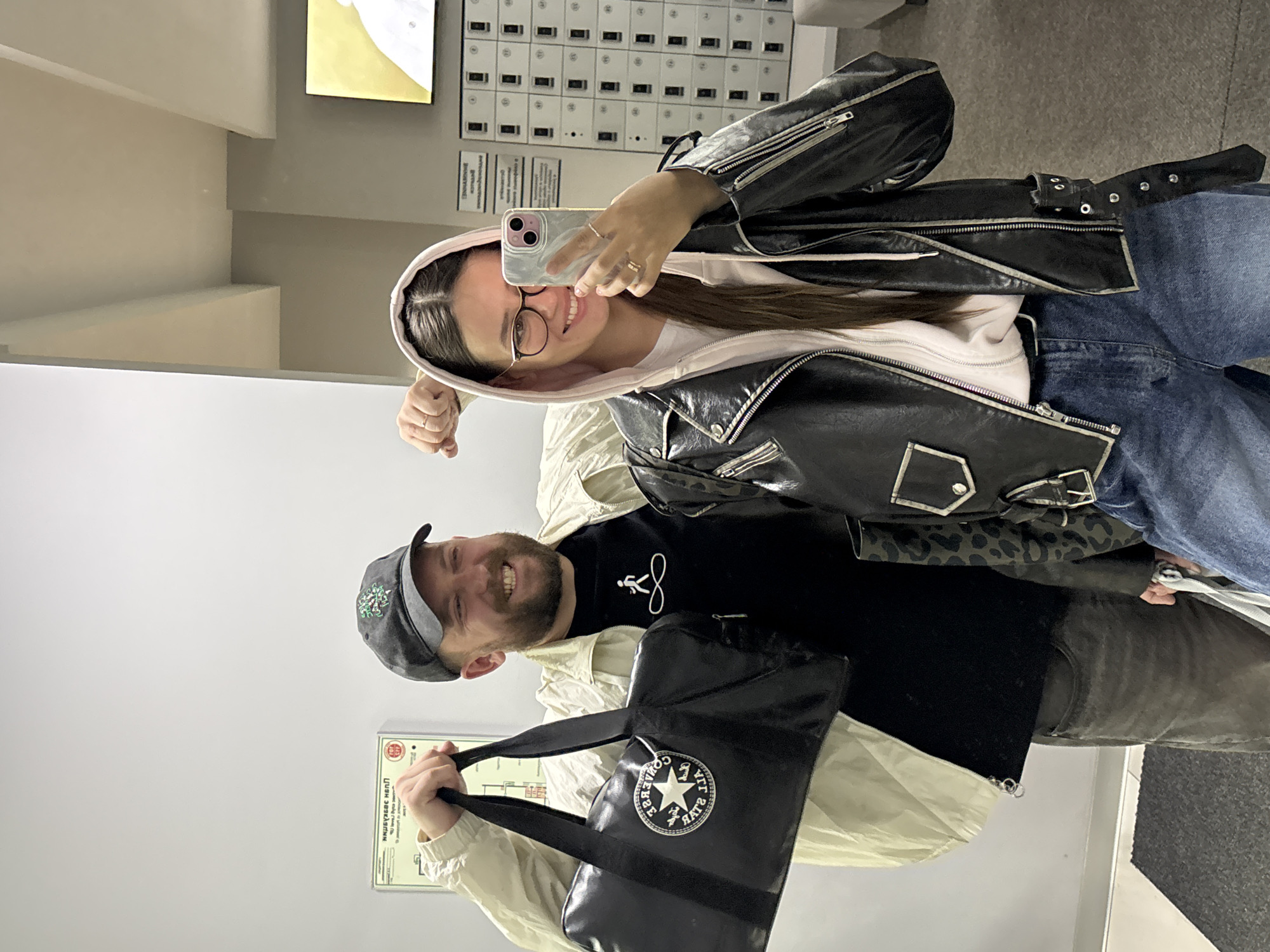A selfie captured indoors in what appears to be a locker room or storage area. Two individuals are visible in the frame. On the top is a woman wearing glasses, a light pink hooded sweatshirt, and a black leather jacket. She is holding the phone up to take the selfie and smiling. The lower figure is a man wearing a baseball cap, a black t-shirt, and a light-colored jacket. He is holding a black Converse branded bag. Lockers are in the background. Based on the sign visible, the location is Krasnoyarsk, Russia.