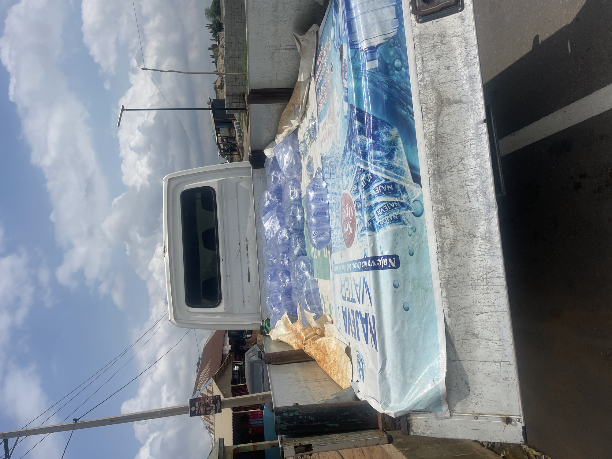 The image shows a white truck carrying a load of bottled water. The truck is parked on a dirt road next to a building. The sky is blue with some white clouds. The bottles of water are arranged in rows and are wrapped in plastic. A large banner with the words "NAJEVA WATER" is draped over the water bottles. The banner has images of water droplets and waves. The truck appears to be in a rural or semi-urban area, possibly in Tasha, Nigeria, given the presence of traditional-style buildings and overhead wires. The lighting suggests it is daytime, likely in the morning or afternoon. The scene seems to be a commercial delivery or transport of goods. There is no visible activity of people, only the parked truck and its cargo.