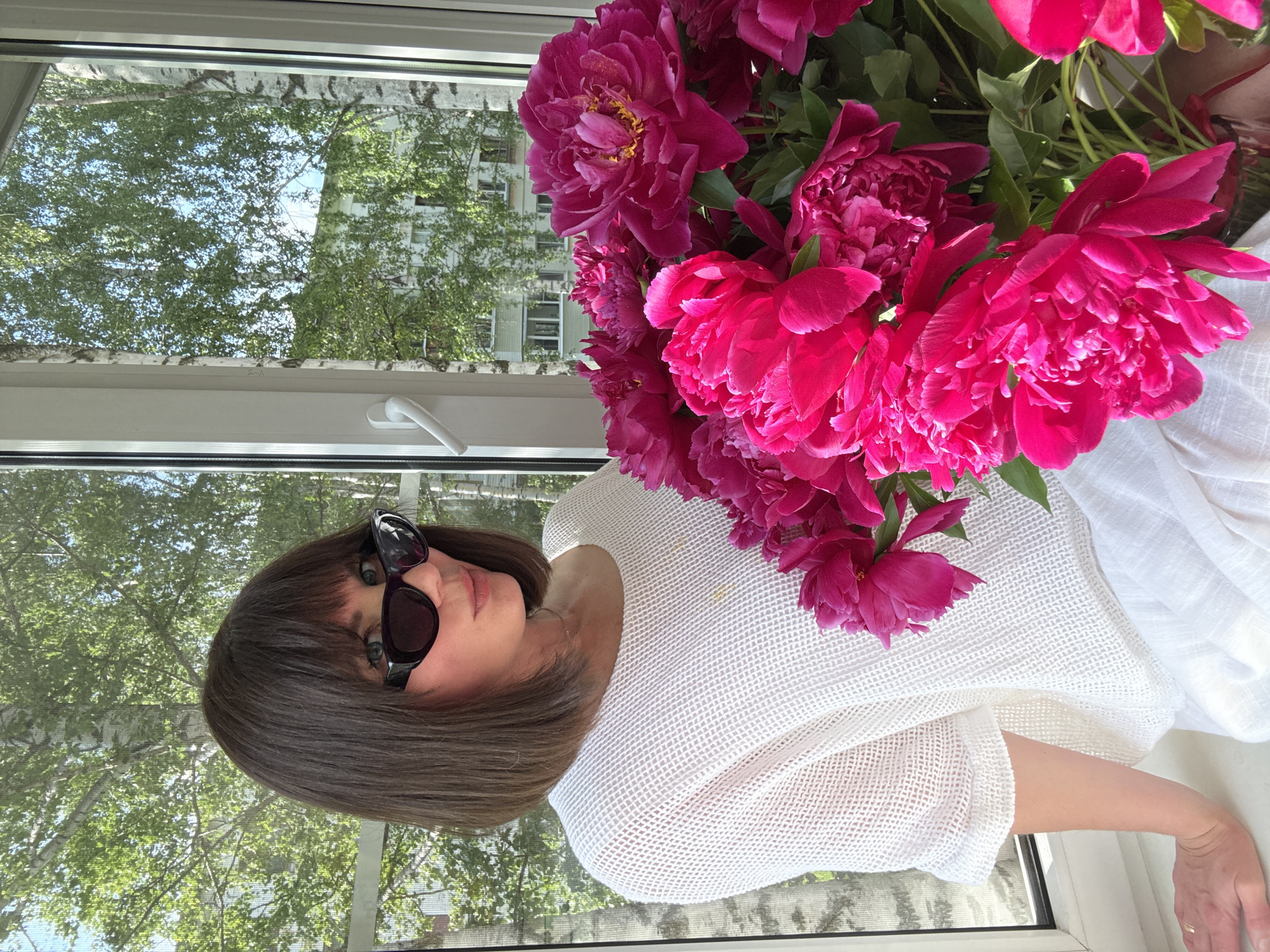 A medium shot captured indoors shows a woman with shoulder-length dark hair and bangs, wearing black cat-eye sunglasses and a white, textured knit top. She is seated on a white windowsill, turned to her right, with a bouquet of vibrant fuchsia peonies to her right. Her left arm rests on the windowsill, and her hand is visible. Behind her, a window overlooks a residential area with buildings and mature trees, likely in the city of Chaikovsky, Russia, given the characteristic birch trees visible. The lighting suggests it is daytime, with sunlight illuminating the scene.