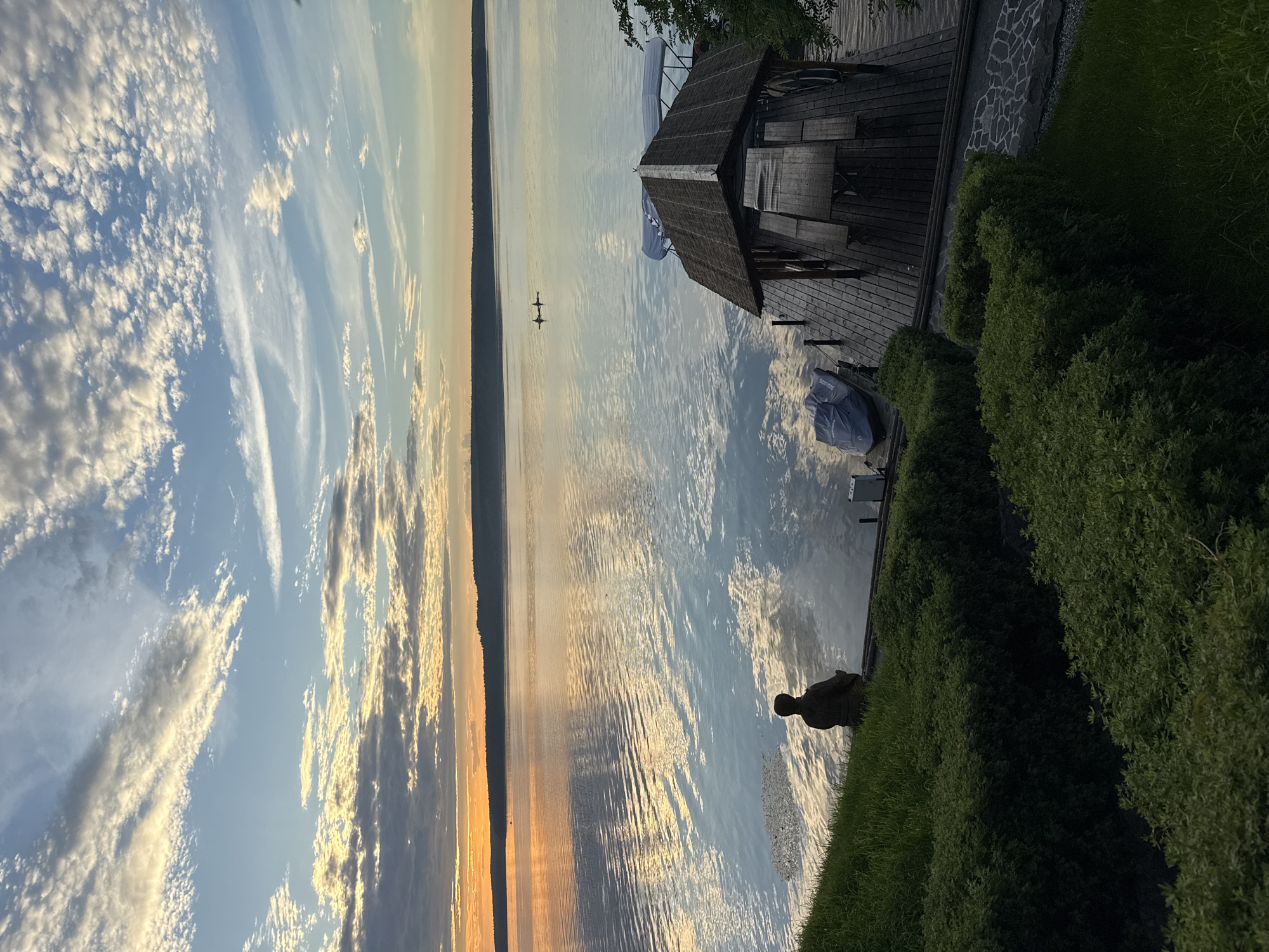 This is an eye-level, vertical shot of a serene lake scene at sunset, likely in Votkinsk, Russia, known for its beautiful waters. In the foreground, a person is seated on the edge of a wooden dock, facing the vast expanse of the lake. The dock is furnished with a covered picnic area, complete with a table and benches. Adjacent to the dock, a jet ski is partially covered by a blue tarp. Lush green bushes and manicured lawns form the immediate surroundings on the left side of the image, sloping down towards the water.
The lake itself is calm, reflecting the soft hues of the sunset and the scattered clouds. In the distance, a dark, tree-lined horizon stretches across the image. Two figures in a kayak are visible on the water, adding a sense of quiet activity to the tranquil landscape. The sky is a canvas of blues and whites, with streaks of golden light from the setting sun illuminating the clouds. The overall atmosphere is peaceful and contemplative.