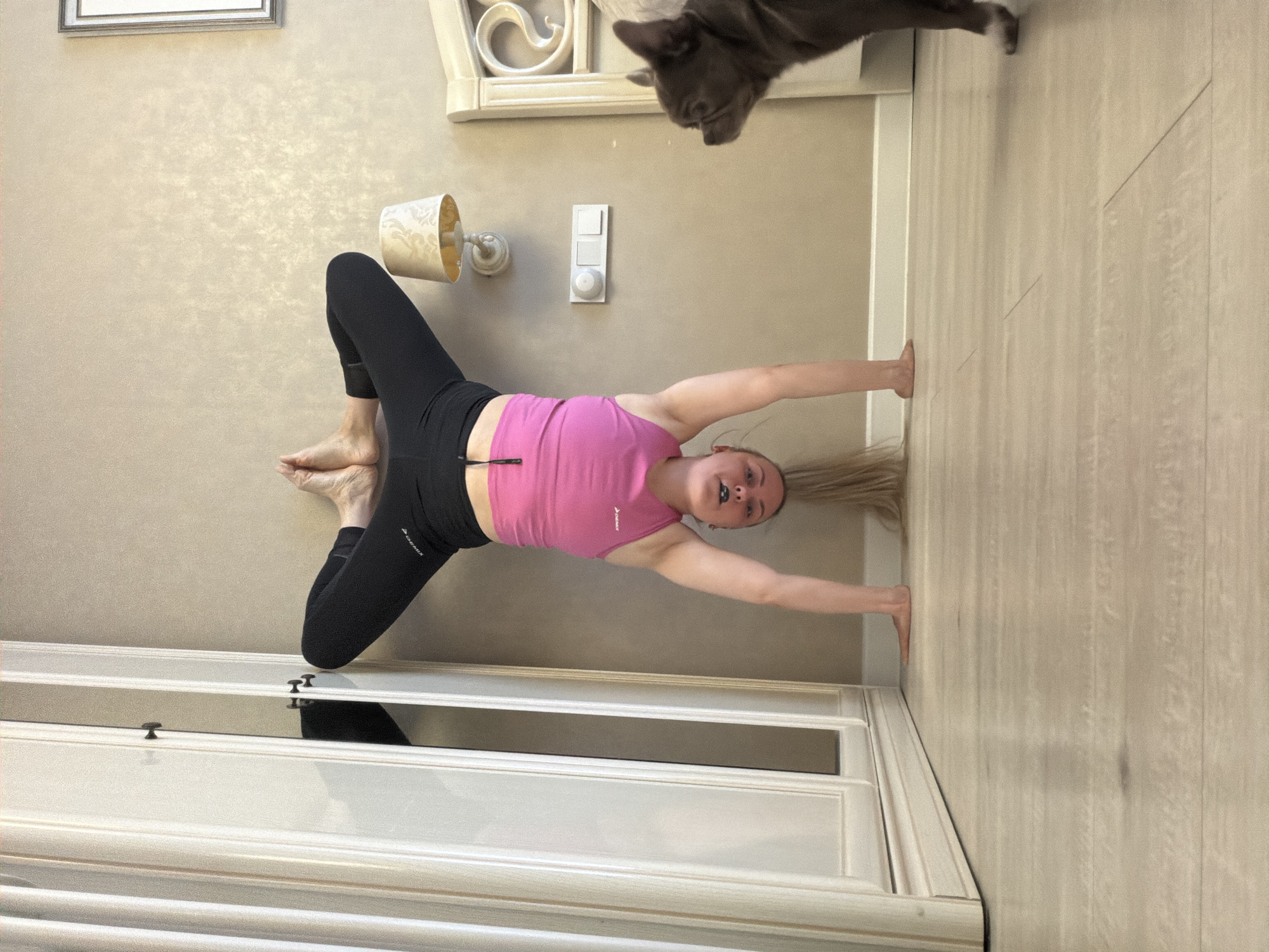 A rotated indoor portrait captures a young woman performing a stretching or yoga pose, observed by a cat. The image is rotated 90 degrees clockwise, making the woman appear to be on a vertical surface, though she is lying on a light wooden floor.
The woman, with long blonde hair spread on the floor, is wearing a pink sports crop top and black leggings with "DEMIX" visible on them. She has a septum piercing. Her legs are bent at the knees, feet flat on the floor, and her arms are extended upwards, parallel to her body. She looks towards the camera with a neutral expression.
In the upper right corner, a dark grey cat, resembling a French Bulldog, peeks down from what appears to be a shelf or piece of furniture. It is looking directly at the woman.
The room features light beige walls with a subtle textured pattern. A wall-mounted lamp with an ornate, light-colored shade is visible on the left. Below it, a white light switch or dimmer is affixed to the wall. A framed picture is partially visible on the wall in the upper left. A white cabinet or dresser with dark handles and decorative molding is seen in the foreground, partially reflecting the room. The overall lighting suggests daytime. The setting is likely a home in Votkinsk, Russia.