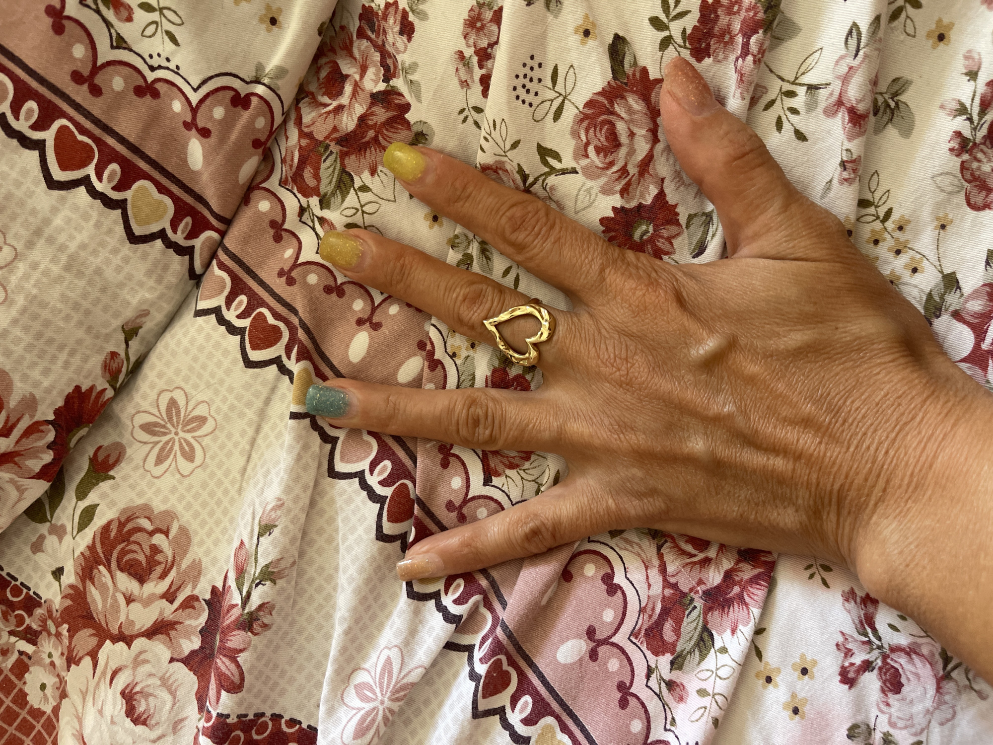 A close-up, overhead shot shows a person's left hand resting on a floral-patterned fabric. The hand is positioned centrally with fingers spread, revealing painted fingernails in various colors: the thumb is painted with a light gold glitter, the index finger a pale gold, the middle finger a light blue-green glitter, and the ring and pinky fingers a bright yellow. A gold heart-shaped ring is worn on the ring finger. The fabric beneath the hand is a white textile adorned with large, stylized red and pink roses, interspersed with smaller yellow flowers and green leaves. A decorative border with scalloped edges and reddish-brown accents runs along the top and right side of the frame. The scene is well-lit, likely indoors. The image does not contain any contextual information about the city of Bosteri, Kyrgyzstan.