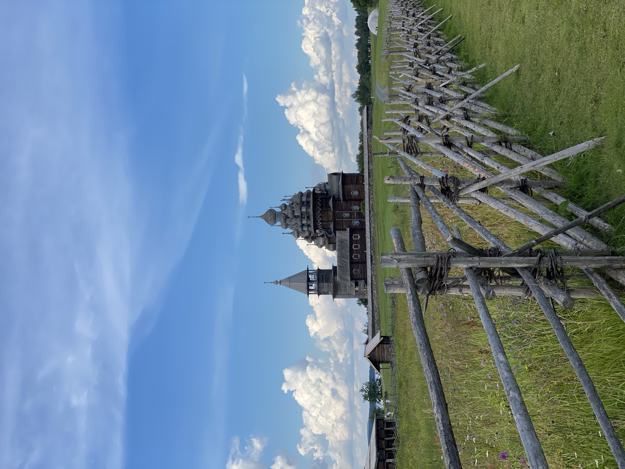 In Boyarshchina, Russia, a historic wooden church with multiple domed roofs and a bell tower stands prominently in a green, grassy field under a blue sky with scattered white clouds. A rustic wooden fence made of logs and branches runs diagonally across the foreground, separating the viewer from the church and a smaller wooden structure to its left. In the background, a body of water is visible, with trees and more distant structures lining the horizon. The scene is tranquil and appears to be a preserved historical site.