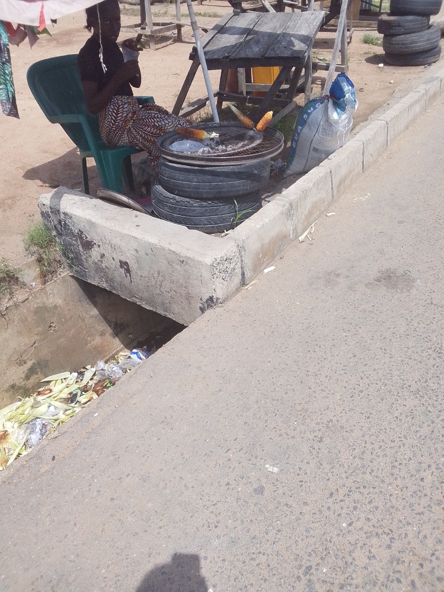 In Jalingo, Nigeria, a woman sits on a green plastic chair in the shade of a makeshift canopy. She is wearing a dark top and a patterned skirt. In front of her, a grill made from tires is cooking corn. To her right, a large bag, possibly of sugar, is propped up against a concrete curb. The scene appears to be a street vendor's stall, with a concrete drain filled with trash to the left of the woman. The ground is a mix of sand and asphalt. The image captures a snapshot of daily life and commerce in the city.