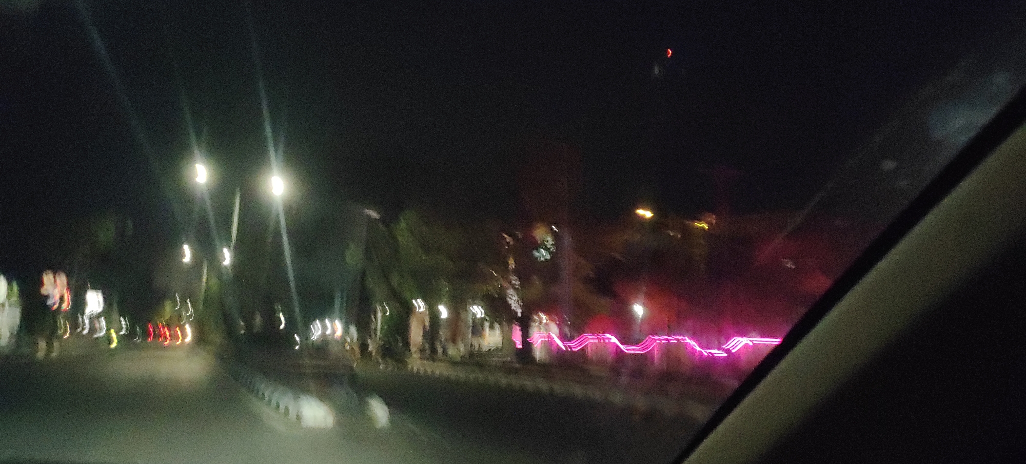 The image captures a nighttime scene on a road in Demak, Indonesia, likely viewed from a moving vehicle. Streetlights illuminate the road and the surrounding trees, casting long streaks of light due to the motion blur. Several brightly lit pink decorative lights are visible along the side of the road, forming a wavy pattern. In the distance, faint red and yellow lights suggest other vehicles or buildings. The overall atmosphere is dark and somewhat hazy, with the blur obscuring finer details of the environment and any potential individuals present.