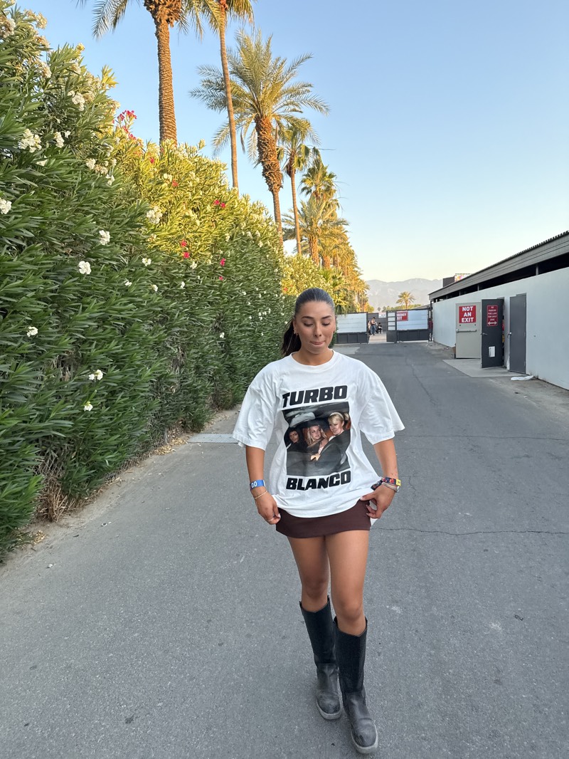 A young woman stands on a paved road in Indio, United States, with a line of trees and foliage to her left and building structures to her right. The woman wears a white t-shirt with a "Turbo Blanco" graphic and a short brown skirt, complemented by knee-high black boots. In the background are tall palm trees and distant mountains under a clear sky. There's a "Not an Exit" sign visible on a building structure in the distance.