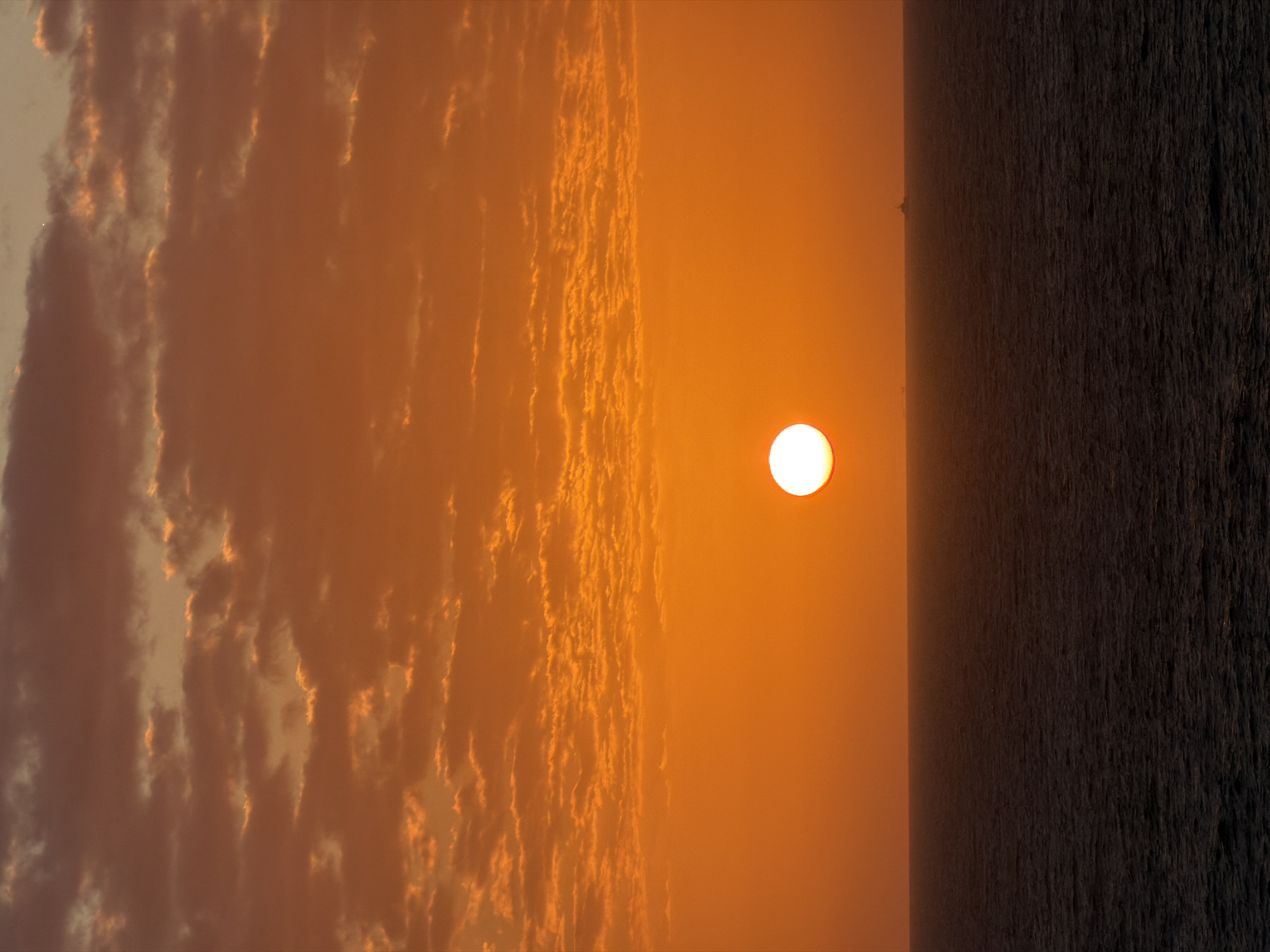 The image depicts a sunset over the ocean. A bright, round sun is positioned slightly above the horizon line. The sky is a gradient of orange hues, transitioning from light near the sun to deeper shades further away. Clouds stretch vertically across the left side of the frame, illuminated by the setting sun. The ocean occupies the right portion of the image, its surface dark and textured. The location is unknown.