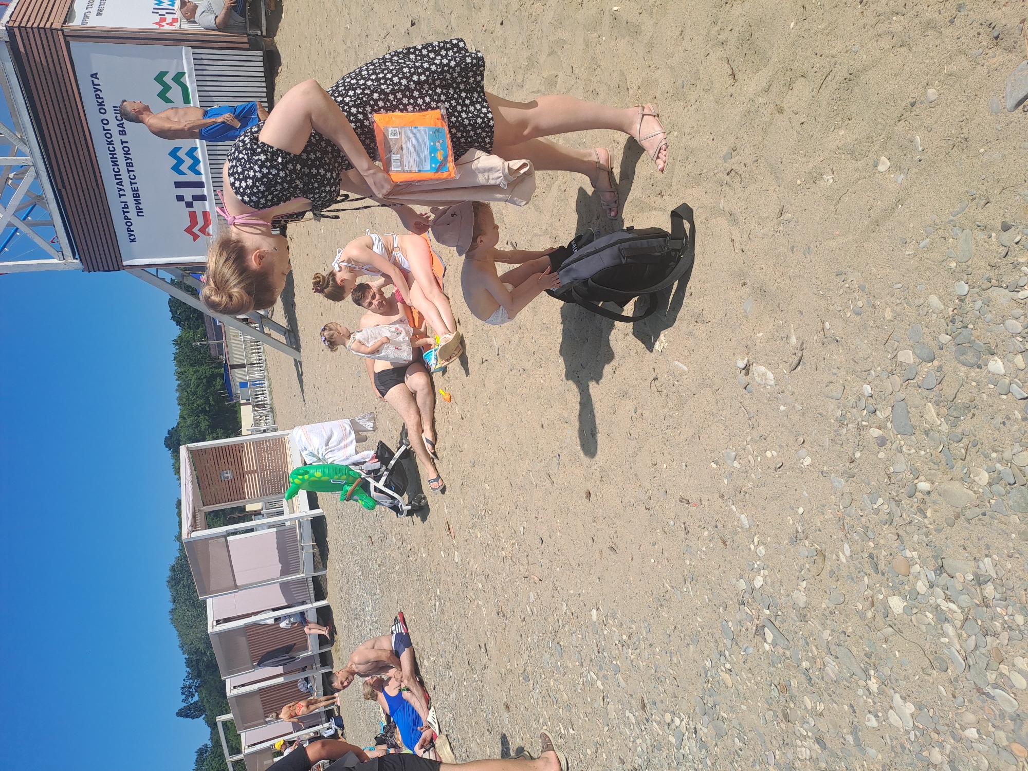 A group of people are on a sandy and pebbly beach under a clear blue sky. In the foreground, a woman in a black floral dress is bending down, holding a beige garment and an orange flotation device. A child wearing a hat and a diaper is also bending down near a black backpack. Behind them, a couple and a young child are relaxing on the sand, with the man and woman lying down and the child standing. A green inflatable dinosaur and a stroller are visible nearby. In the background, there are rows of white beach cabanas and a lifeguard tower with signage in Russian that reads "KURORTY TUAPSE DISTRICT WELCOMES YOU." Several other people are scattered on the beach and near the cabanas. The scene appears to be a sunny day at a resort in Dzhubga, Russia.