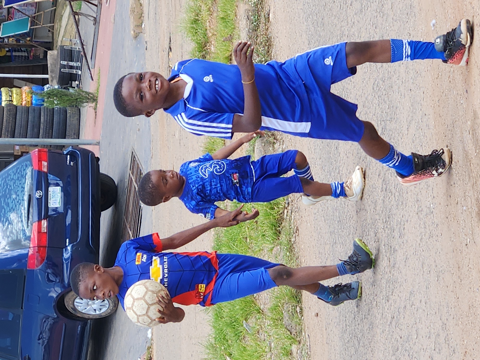 The image depicts three young boys in blue soccer uniforms walking on a dirt road in Jos, Nigeria. The boy in the foreground on the right is smiling and running, wearing a royal blue jersey with white stripes on the sleeves and white shorts. He has on blue socks and black and pink soccer cleats. The boy in the middle is walking, holding the hand of the boy on the left. He wears a blue jersey with a white number 3 on it and blue shorts. The boy on the left is holding a well-worn soccer ball in his right arm. He wears a blue jersey with red accents, a yellow Chevrolet logo, and blue shorts. He is wearing blue socks and dark grey soccer cleats with lime green soles. In the background, a dark blue minivan is parked. Further in the background, there are stacks of tires, a small tree, and various market stalls. The ground is a mix of dirt and gravel, with some sparse green grass growing along the sides. The overall scene suggests a casual outdoor activity, possibly a game of soccer, in a common urban environment in Jos.