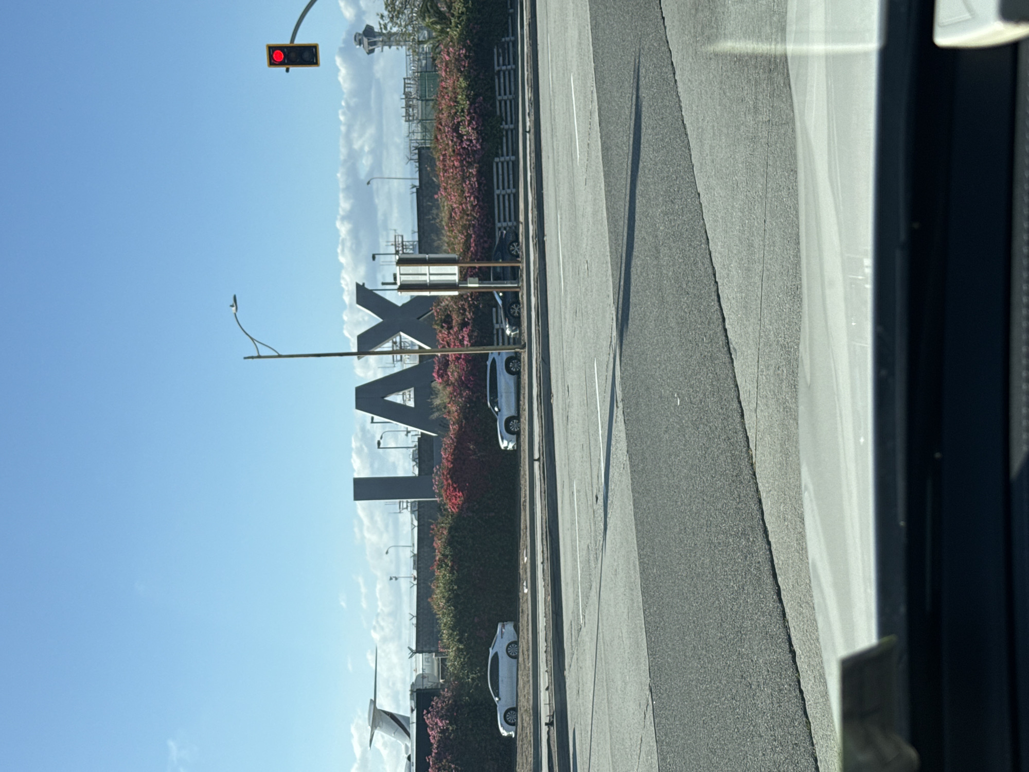 The image shows the iconic "LAX" sign located in Los Angeles, California. The sign is situated on the side of the road, adjacent to a flower hedge. There are some cars parked alongside the road next to the sign. A red traffic light is visible in the distance. The sky is clear with some clouds. The image appears to be taken from inside a vehicle.