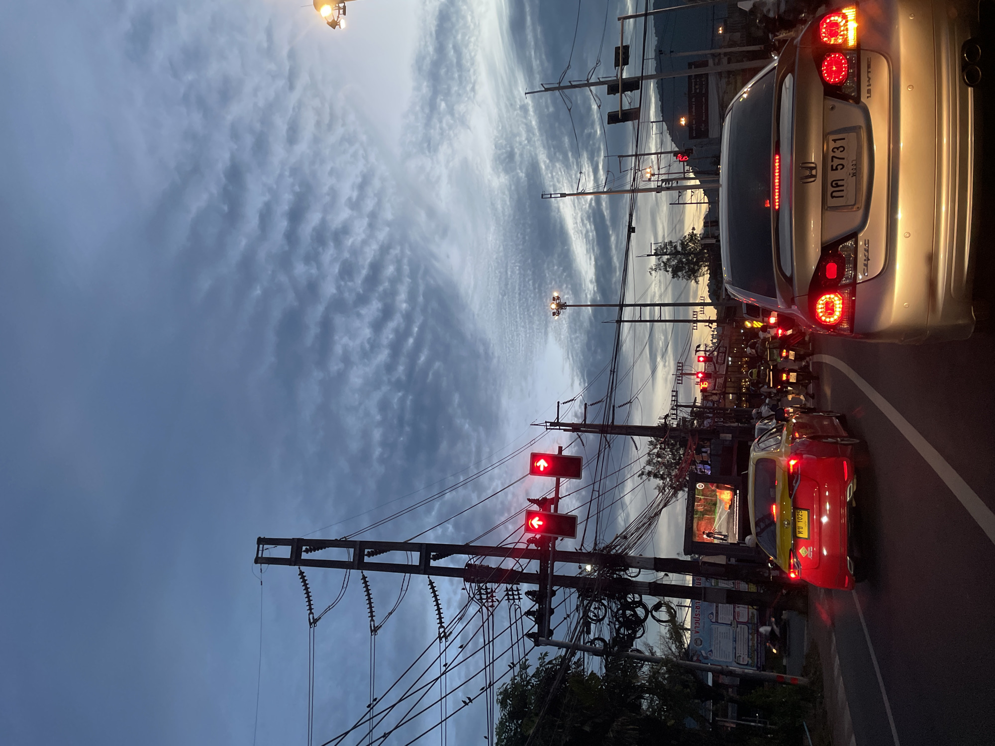 The image depicts a street scene at dusk in an urban environment. The sky is overcast with textured clouds. In the foreground, traffic is stopped at a red light. A silver Honda Civic, bearing the license plate "กค 5731," is prominently featured, its brake lights illuminated. To its left, a red car, likely a taxi, with a yellow roof and the license plate "พษ 1025," is also stopped. A multitude of electrical wires and poles dominate the midground, with traffic lights showing red for the direction of travel. In the background, streetlights, more traffic signals, and the outlines of buildings and hills are visible. A large digital billboard displays an image. The overall atmosphere suggests a busy intersection during evening commute hours.