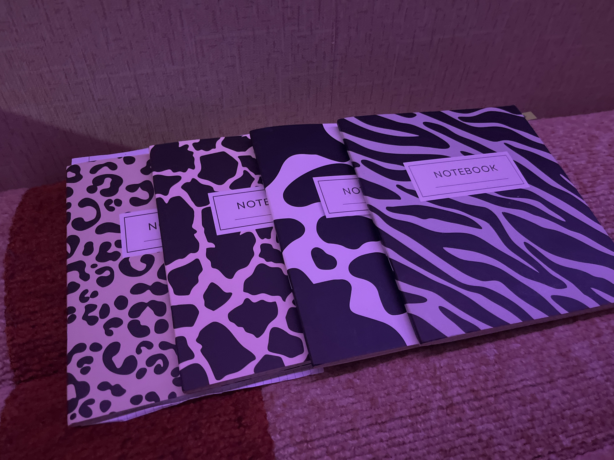 The image shows four notebooks stacked on top of each other on a textured surface. The notebooks have different animal print patterns: leopard, cow, and zebra. Each notebook has a white rectangular label on the cover, with the words "NOTEBOOK" or "NOTE" printed on them. The background is blurred and appears to be a textured wall or surface. The scene is lit with a purple hue. There is no information to indicate the location is in the city of Chaikovsky, Russia.