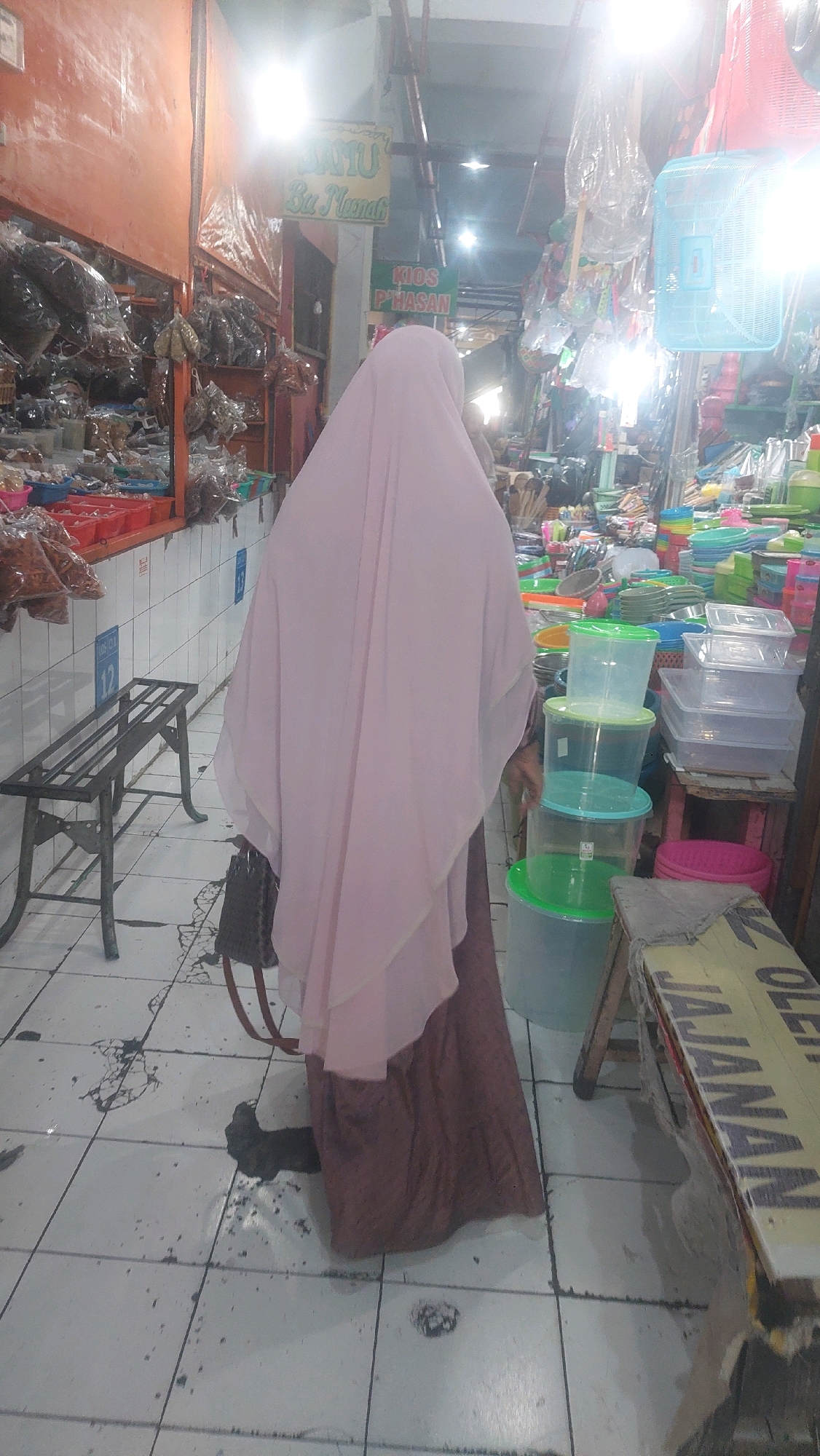 A person wearing a light pink hijab and a brown dress stands in a busy marketplace in Madiun, Indonesia. The person is facing away from the camera, looking into a stall filled with plastic containers of various sizes and colors. In the background, stalls are packed with merchandise, including bags of dried goods and other household items. The floor is tiled with white and black squares, and there are some spills and debris on the ground. A metal bench is visible to the left of the person, and a wooden stall with a yellow sign that reads "JAJANAN" (snacks) is to the right. The overall impression is of a vibrant and crowded market scene.