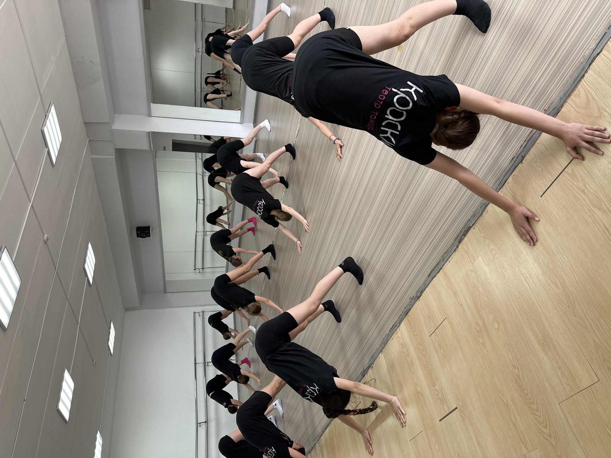 A group of young people, likely children or teenagers, are engaged in a synchronized stretching or dance exercise inside what appears to be a dance studio in Votkinsk, Russia. The room features a light wooden floor with visible seams, a mirrored wall on the left reflecting more participants and a barre, and white walls on the right. The ceiling is equipped with multiple rectangular fluorescent light fixtures, providing artificial illumination.
The participants are dressed uniformly in black short-sleeved t-shirts and black shorts. Most wear black socks, though some have white socks, and a few have pink footwear or are barefoot. They are arranged in several rows, performing a forward-bending pose where they lean forward from the hips, extending their arms towards the floor with their fingers spread, and keeping their legs straight. This posture suggests a focus on flexibility and core strength, typical for dance or gymnastics training.
Visible text on the back of the t-shirts is in Cyrillic script. The prominent text reads "КРОКИ" (KROKI), with smaller text below it stating "ТЕАТР ТАНЦА" (THEATER OF DANCE), indicating they are part of a group named "KROKI DANCE THEATER". The disciplined and synchronized nature of their movements suggests they are participating in an organized class or practice session. The indoor setting and artificial lighting do not provide cues for the time of day or weather.