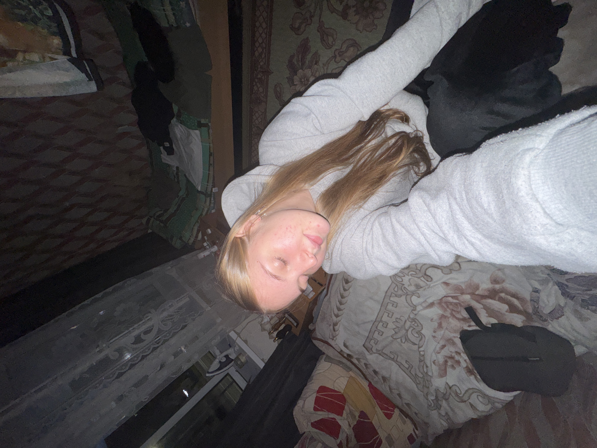 The image shows a young woman lying on a bed in a room. She is wearing a light gray hooded sweatshirt and matching sweatpants. Her blonde hair is partially covering her face. The bed is covered in a patterned comforter with shades of brown, red, and white. A dark-colored pillow or object lies next to her. A window with lace curtains is visible in the background. The room appears to be located in an older building in the city of Perm, Russia.
