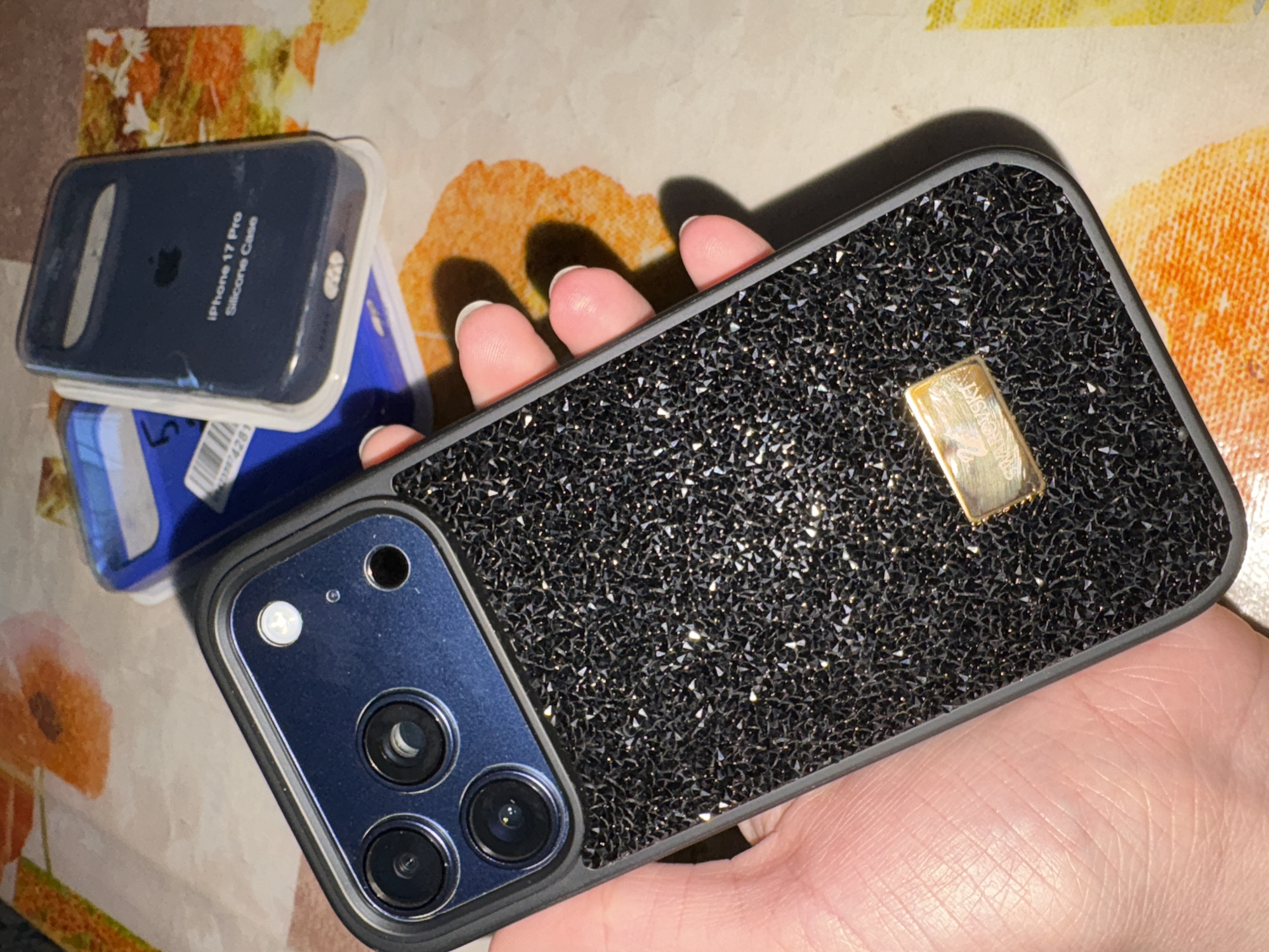 The image shows a hand holding a smartphone in a black, sparkly case with a gold rectangular accent piece. The phone has a triple-lens camera system. To the left of the phone is an "iPhone 17 Pro Silicone Case" still in its packaging, along with a blue plastic card. The background is a surface with an orange and beige floral pattern. There is no indication in this image of the city of Rusalevka, Russia.