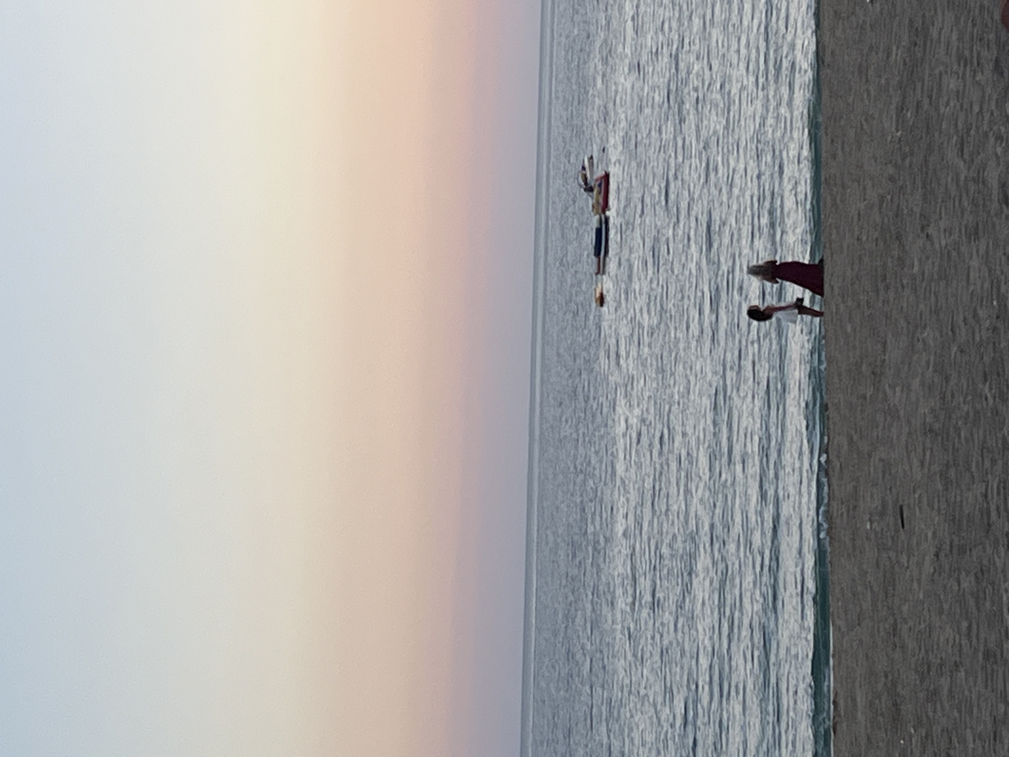 This image captures a tranquil beach scene, likely at either sunrise or sunset, in Alanya, Turkey. The sky occupies the upper portion of the frame, displaying a soft gradient of colors that transition from a pale bluish-grey on the left to a light peachy-pink and orange on the right, indicative of twilight hours. Below the horizon, the expansive sea stretches across, its calm, grey-blue surface subtly textured by small, consistent ripples reflecting the gentle light.
In the water further out, two individuals are visible. One appears to be on an inflatable object, while the other is standing or wading nearby. Closer to the shore, towards the bottom right of the image, two more people are present. One figure, wearing a white top, stands partially immersed in the water, while another person, in a red garment, is partially visible on the dark grey sand of the beach. The sand has a noticeable coarse texture.
The scene depicts individuals enjoying the calm waters and the peaceful ambiance of the coast during either dawn or dusk. There is no visible text in the image.