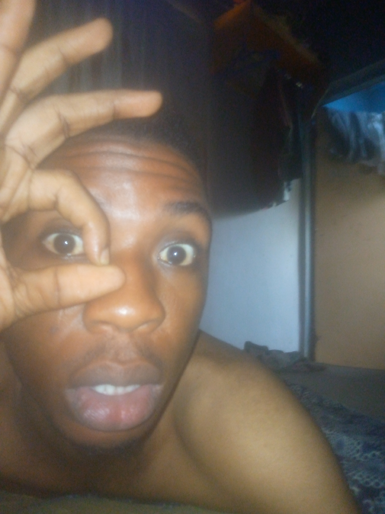 A close-up shot of a dark-skinned, shirtless person making an "OK" sign over their right eye. The person is looking directly into the camera with their mouth slightly open. Behind the person, there is a white wall, a brown door frame, and some indiscernible items hanging overhead. The location appears to be indoors. The city is Unknown, Unknown.