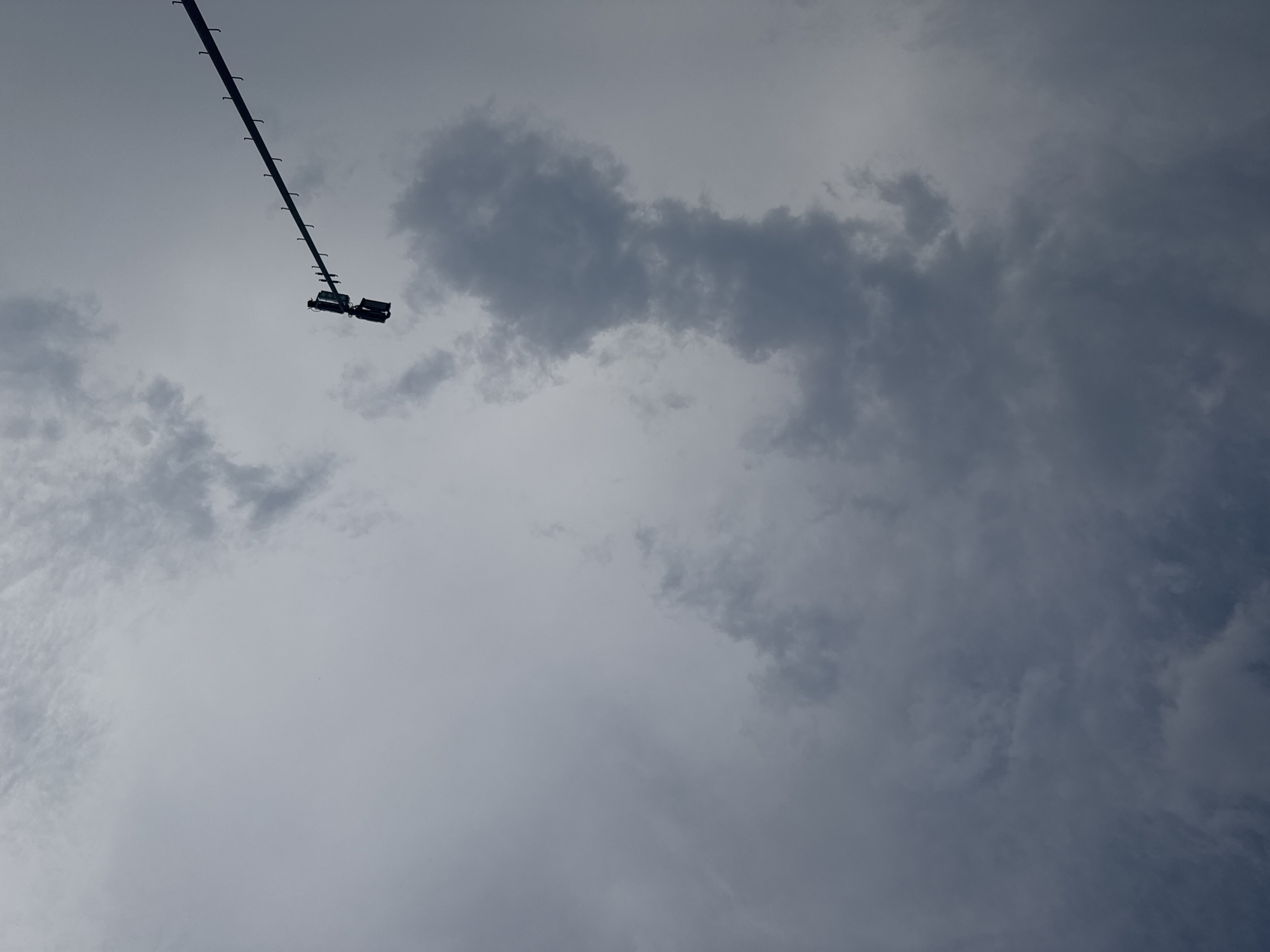The image captures a view looking upwards at a cloudy sky. A traffic light is mounted on a pole that extends diagonally across the upper right portion of the frame. The traffic light appears to have three distinct lenses, with the top two illuminated. The sky is overcast with a mix of light and dark gray clouds. The scene is likely located in Chiasso, Switzerland, given the context.